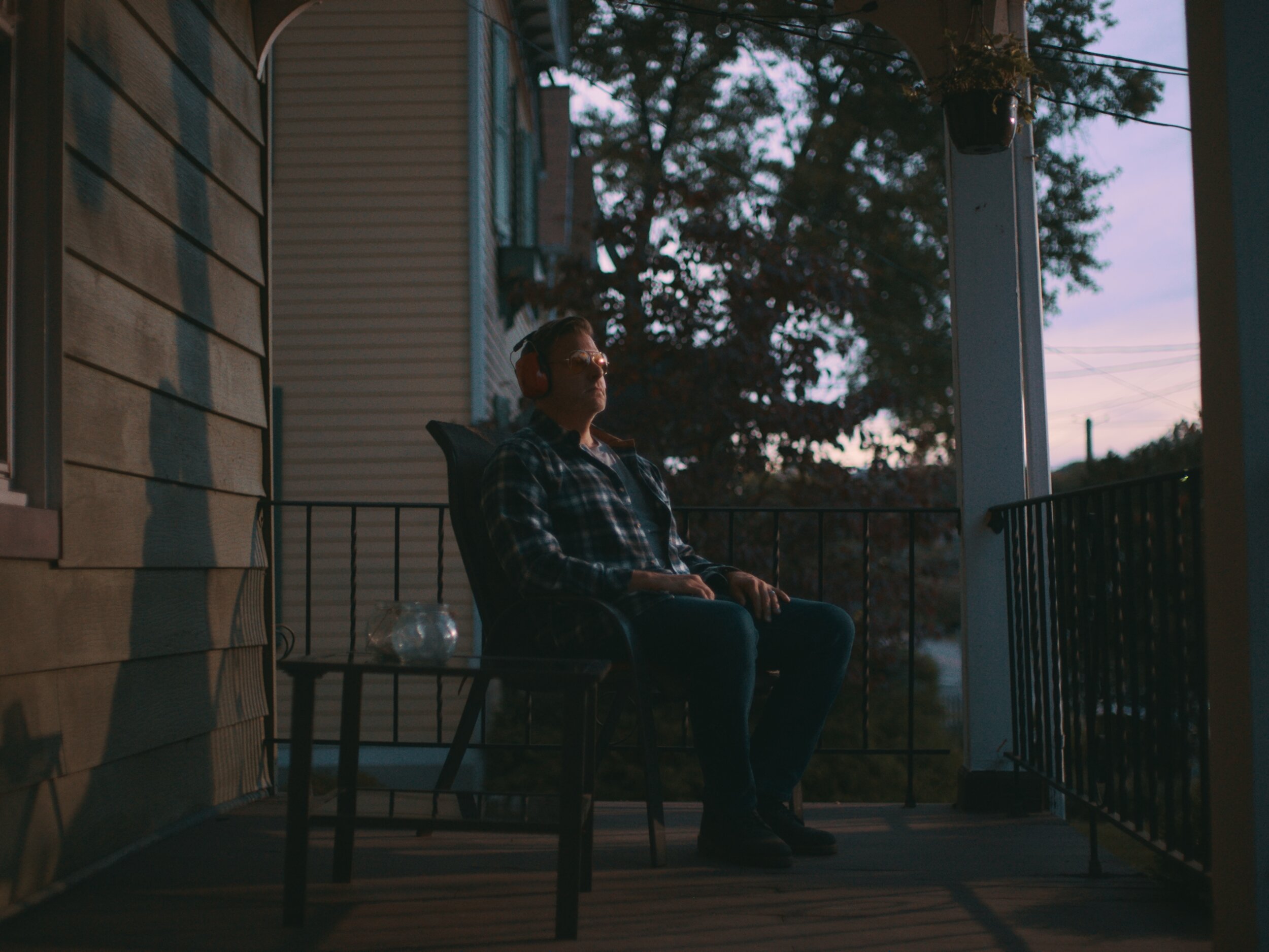 Person sitting outdoors on a porch, wearing headphones and sunglasses, at dusk or dawn.