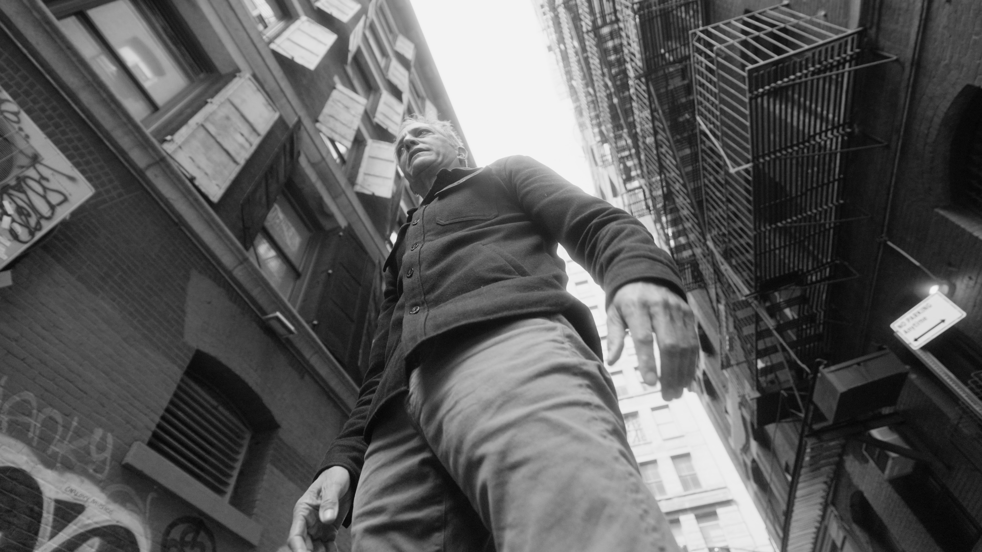 Low-angle black-and-white photo of a man standing on a city street with tall buildings and fire escape stairs around him.