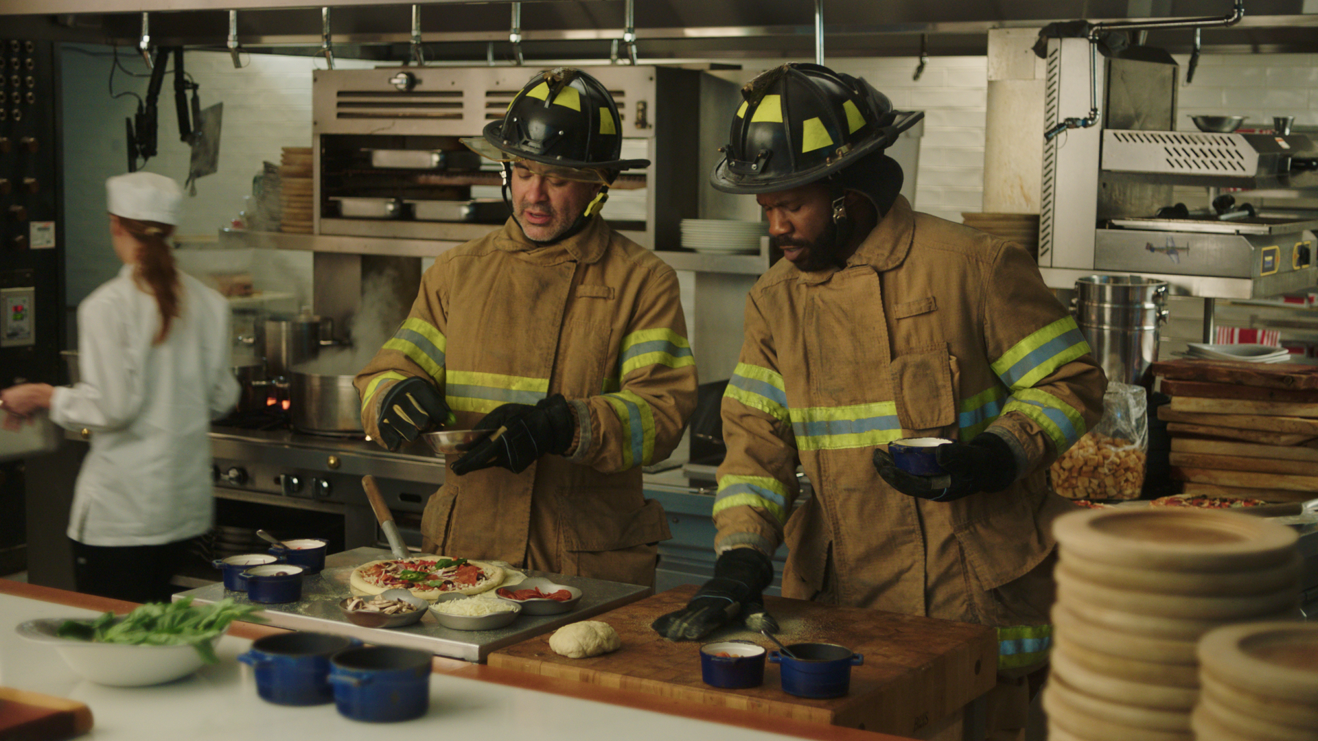 Two firefighters in uniform with helmets preparing food in a commercial kitchen, with a chef in white in the background.