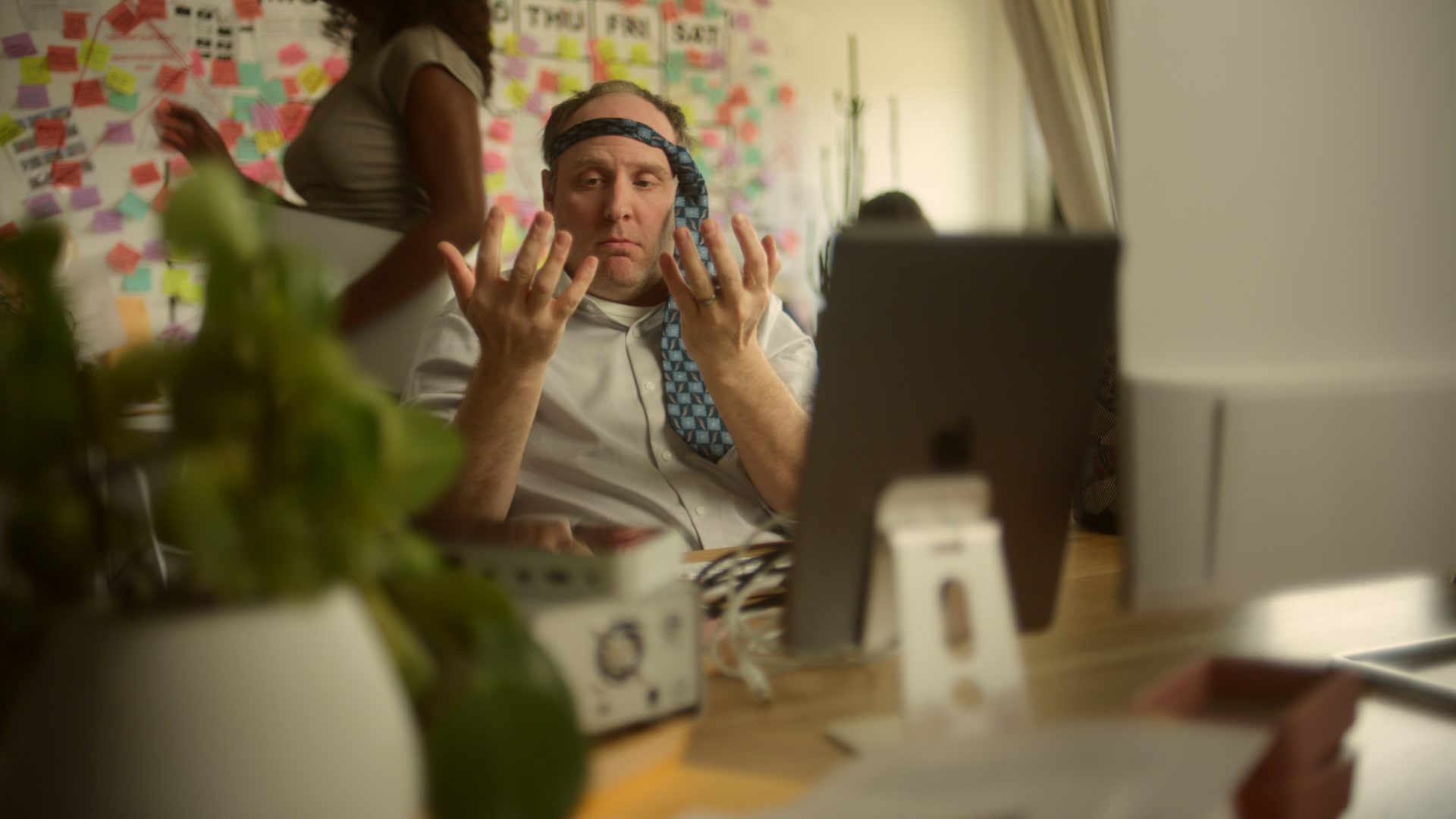 Man with a tie, wearing a headband, sitting at a desk in front of a computer, with a woman in the background and a wall full of colorful sticky notes.