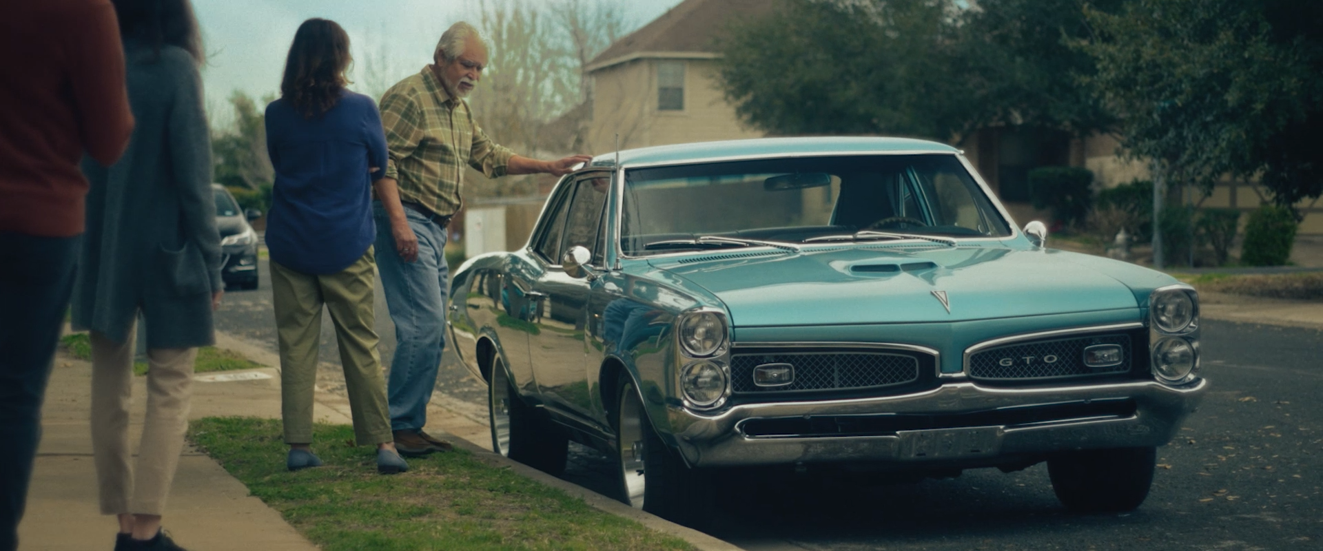 A vintage blue Pontiac GTO parked on a suburban street with four people gathered around it, examining or discussing the car.
