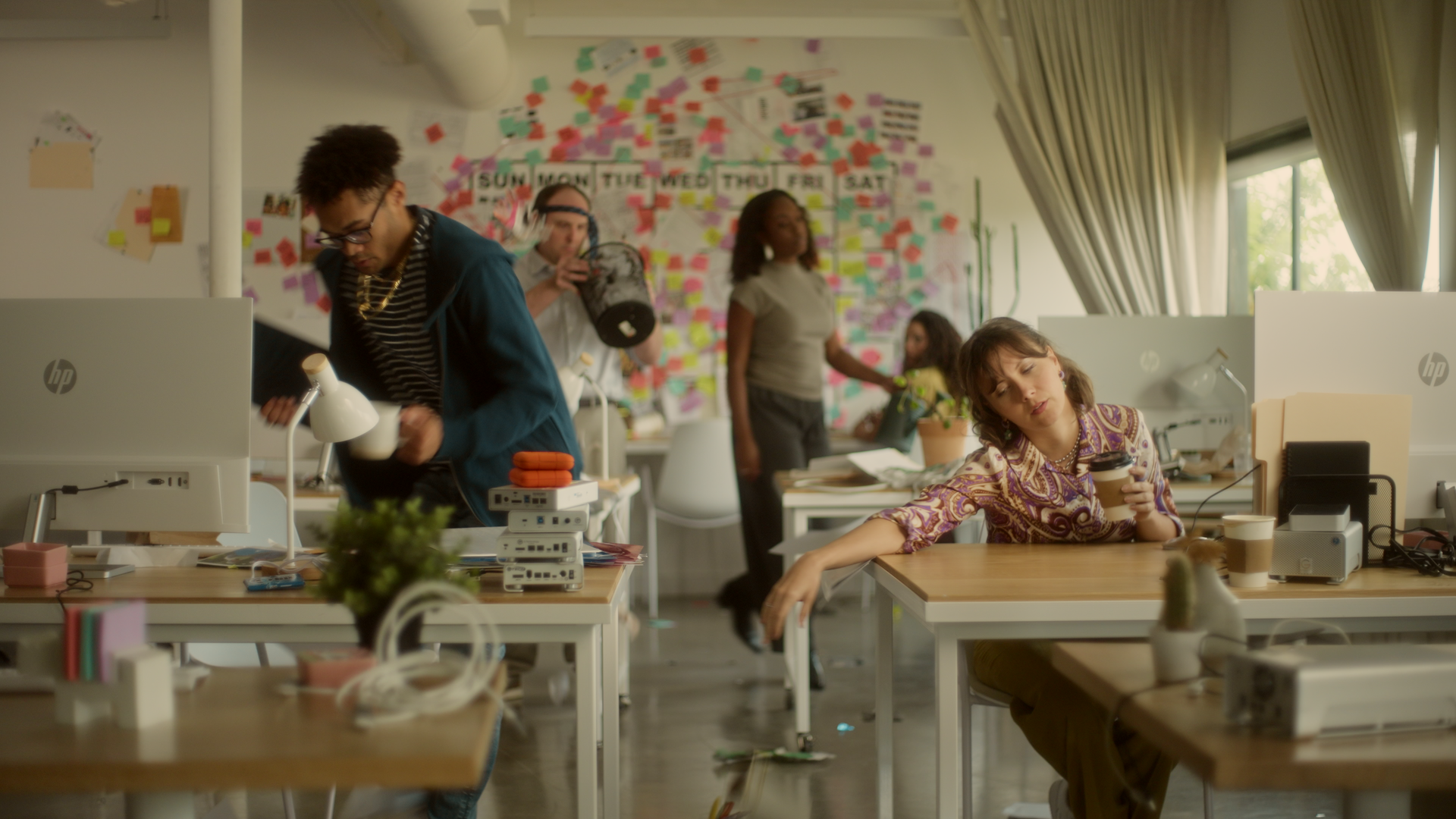 An office scene with multiple people, some working at desks with computers and others walking or sitting, with a wall covered in colorful sticky notes and a large window with curtains.