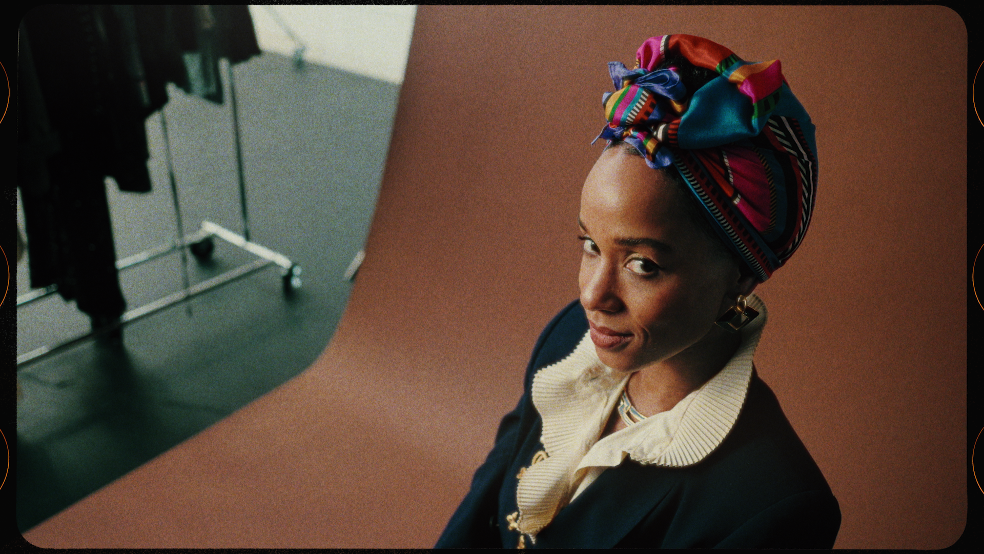 A woman with a colorful headwrap looking up at the camera, sitting on a pink background, with some clothing racks visible in the blurred background.