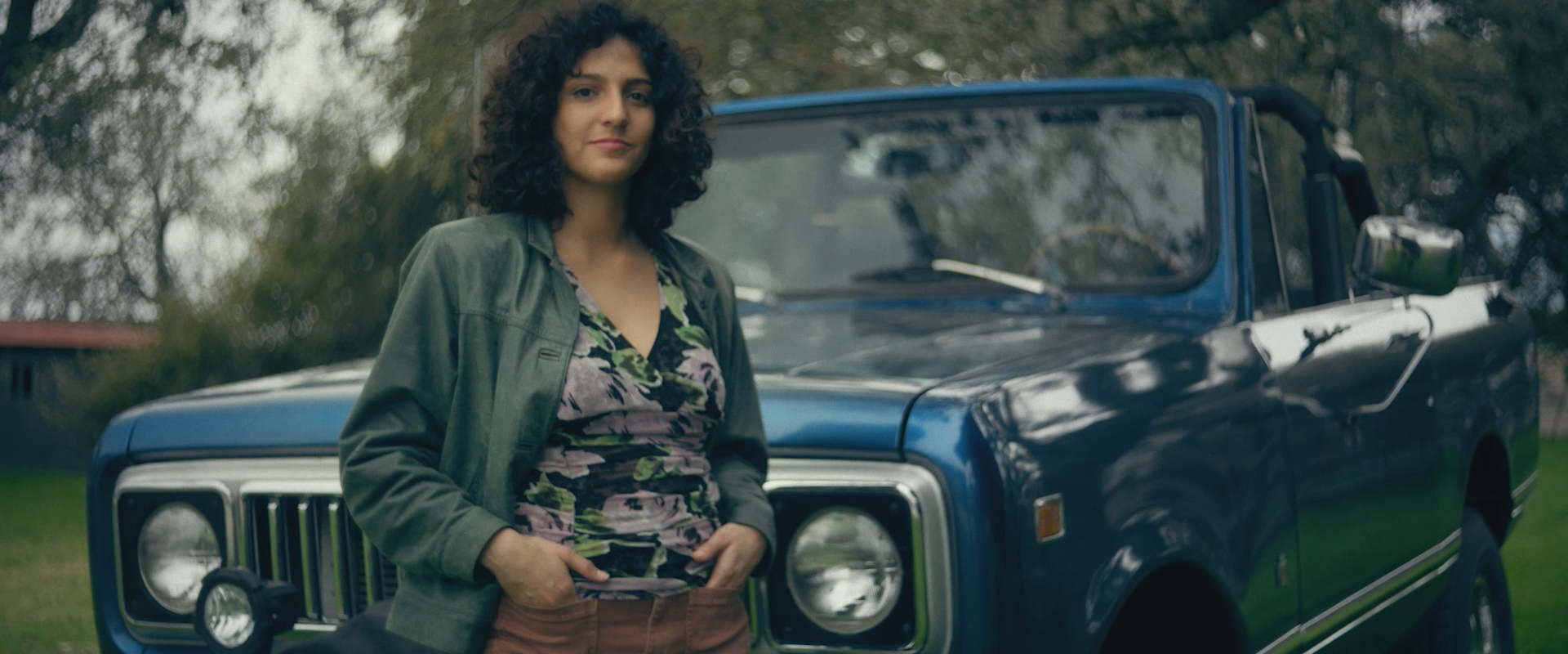 A woman with curly dark hair wearing a green jacket and a floral top standing in front of a blue Jeep parked outdoors with trees in the background.