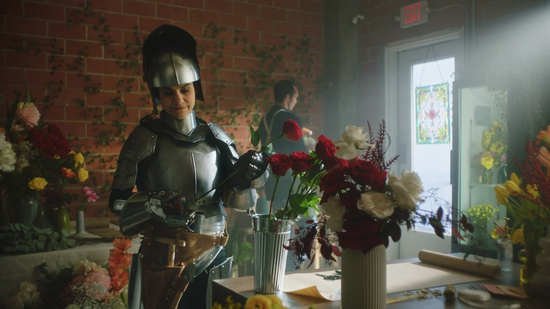 A woman dressed as a medieval knight working at a flower shop with colorful flowers and vases, another person in the background, sunlight streaming in.