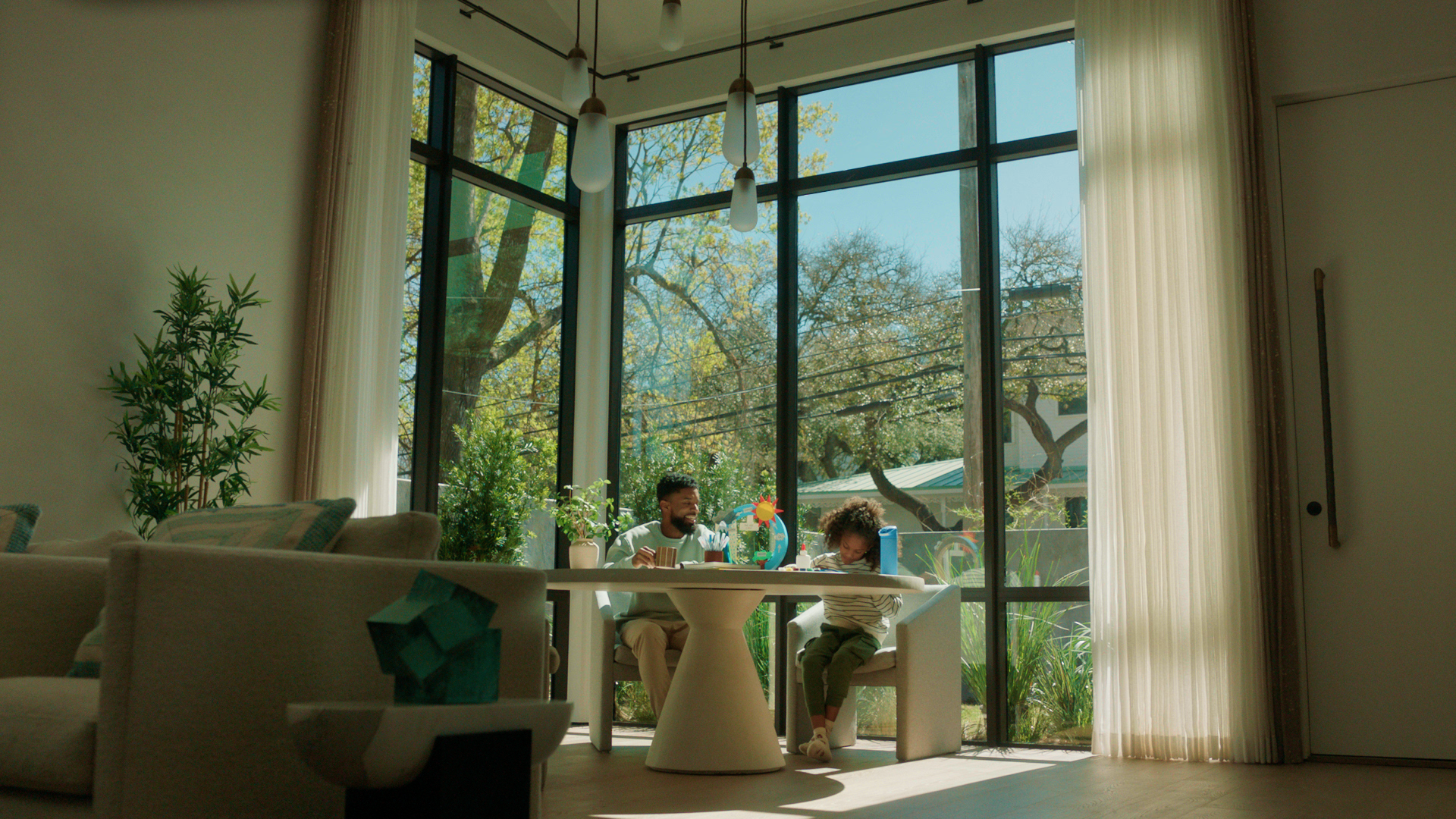 A man and a young girl sitting at a white dining table in a bright room with large glass windows, greenery outside, beige curtains, some houseplants, and modern hanging light fixtures.