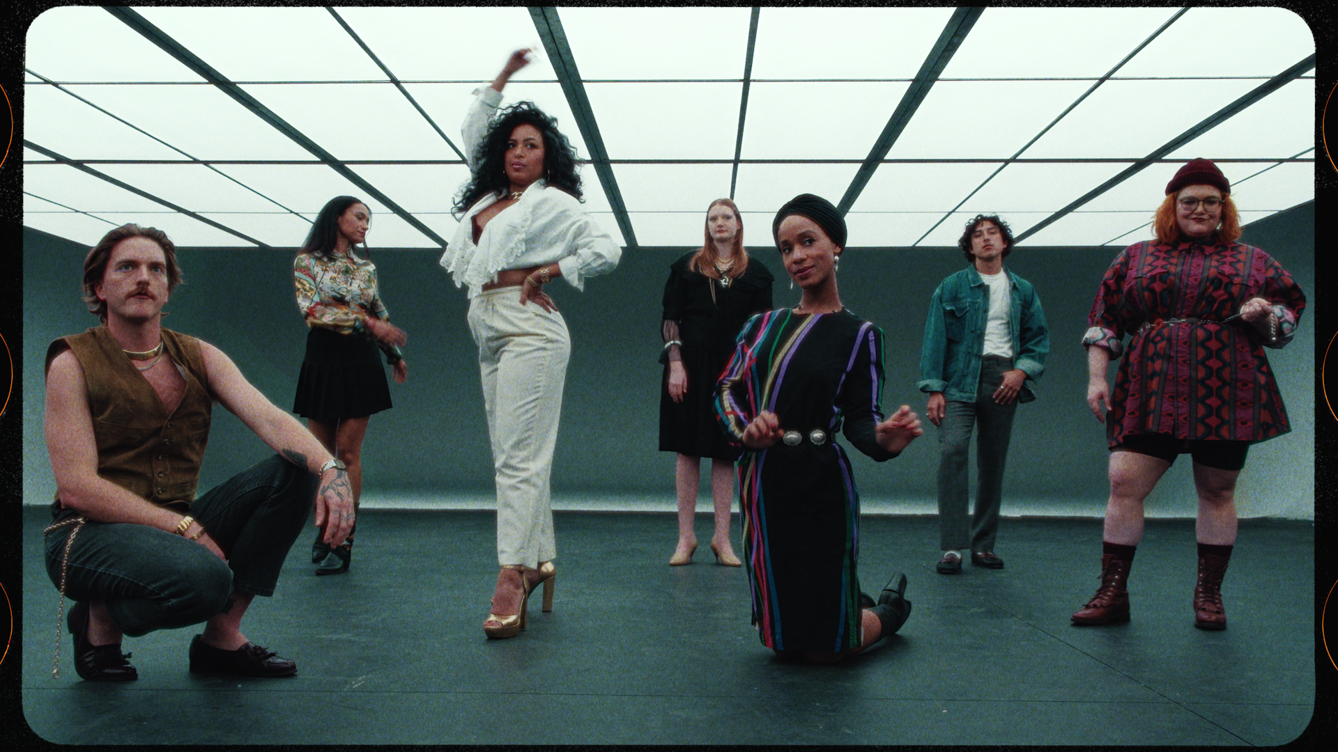 Group of seven diverse individuals in a dance studio with a minimalist ceiling, dressed in retro and eclectic fashion, posing confidently.