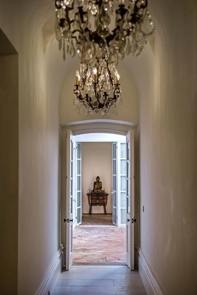 View along a barrel-vaulted whitewashed corridor at Casa Fuzetta lit by a crystal chandelier, through open glazed doors to a gilded seated Buddha on an antique table flanked by candles.