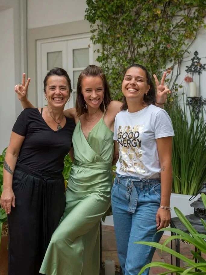 Cheeky Yoga Club founder Kat Achtelik with Casa Fuzetta House Manager Claudia and masseuse Justine, laughing together in a plant-filled corner in Olhão, one wearing a 'Good Energy' t-shirt.