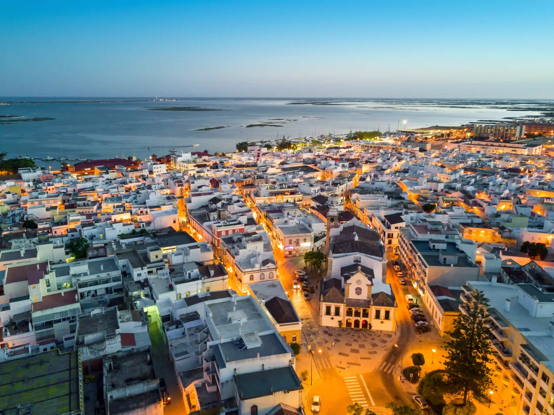 An aerial view of Olhao at dusk, home to Casa Fuzetta; lamplit streets around the Igreja Matriz and whitewashed rooftops running down to the market halls, marina and Ria Formosa islands.