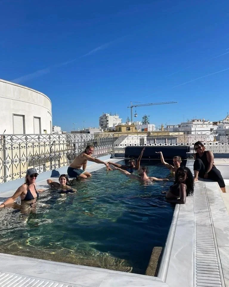 A Cheeky Yoga Club retreat group in the rooftop pool at Casa Fuzetta in Olhão, laughing together with hands raised, whitewashed cubist rooftops beyond the wrought-iron balustrade.