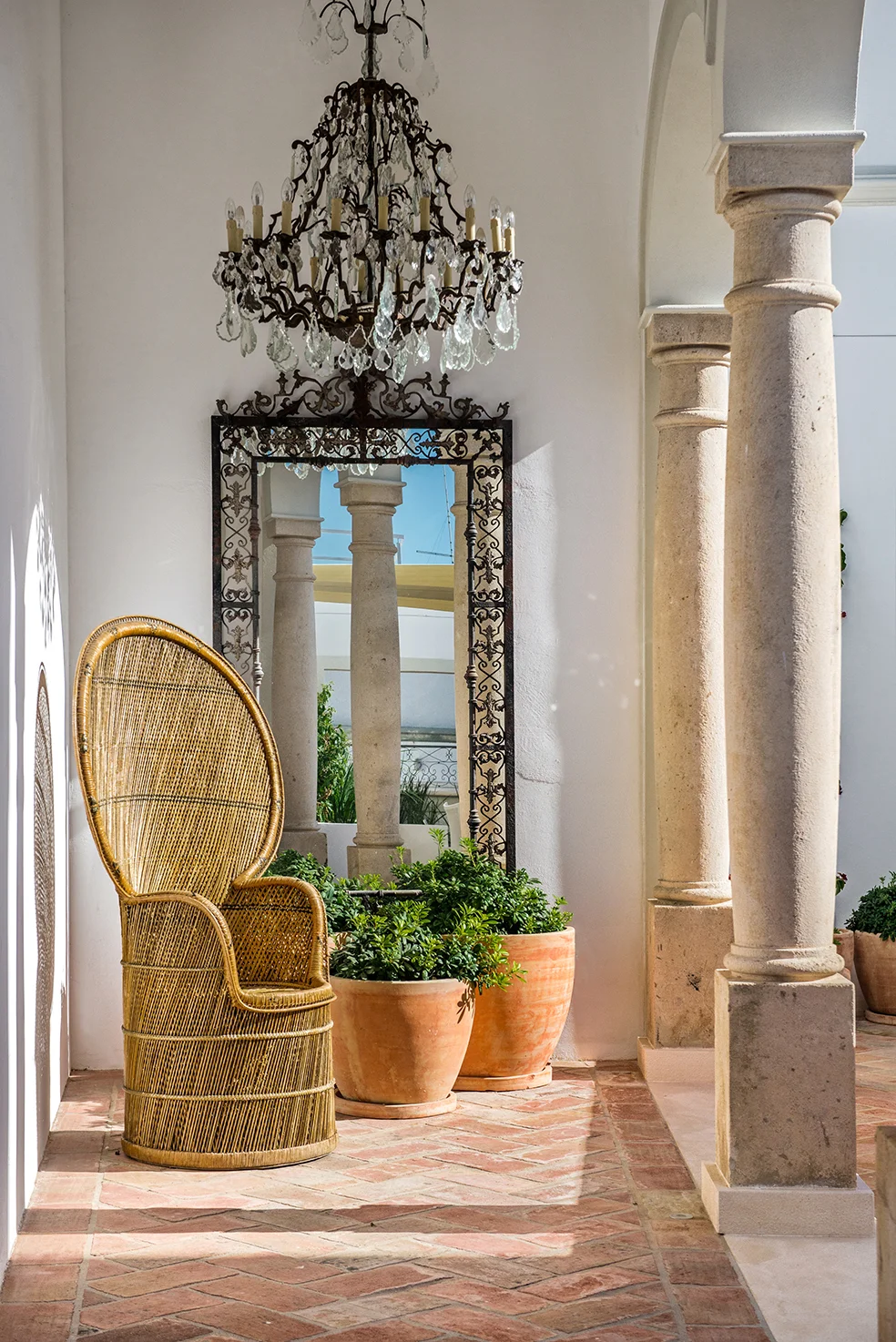 Sunlit whitewashed interior at Casa Fuzetta, Olhão — an antique crystal chandelier and iron-framed mirror beside a peacock rattan chair and terracotta planters