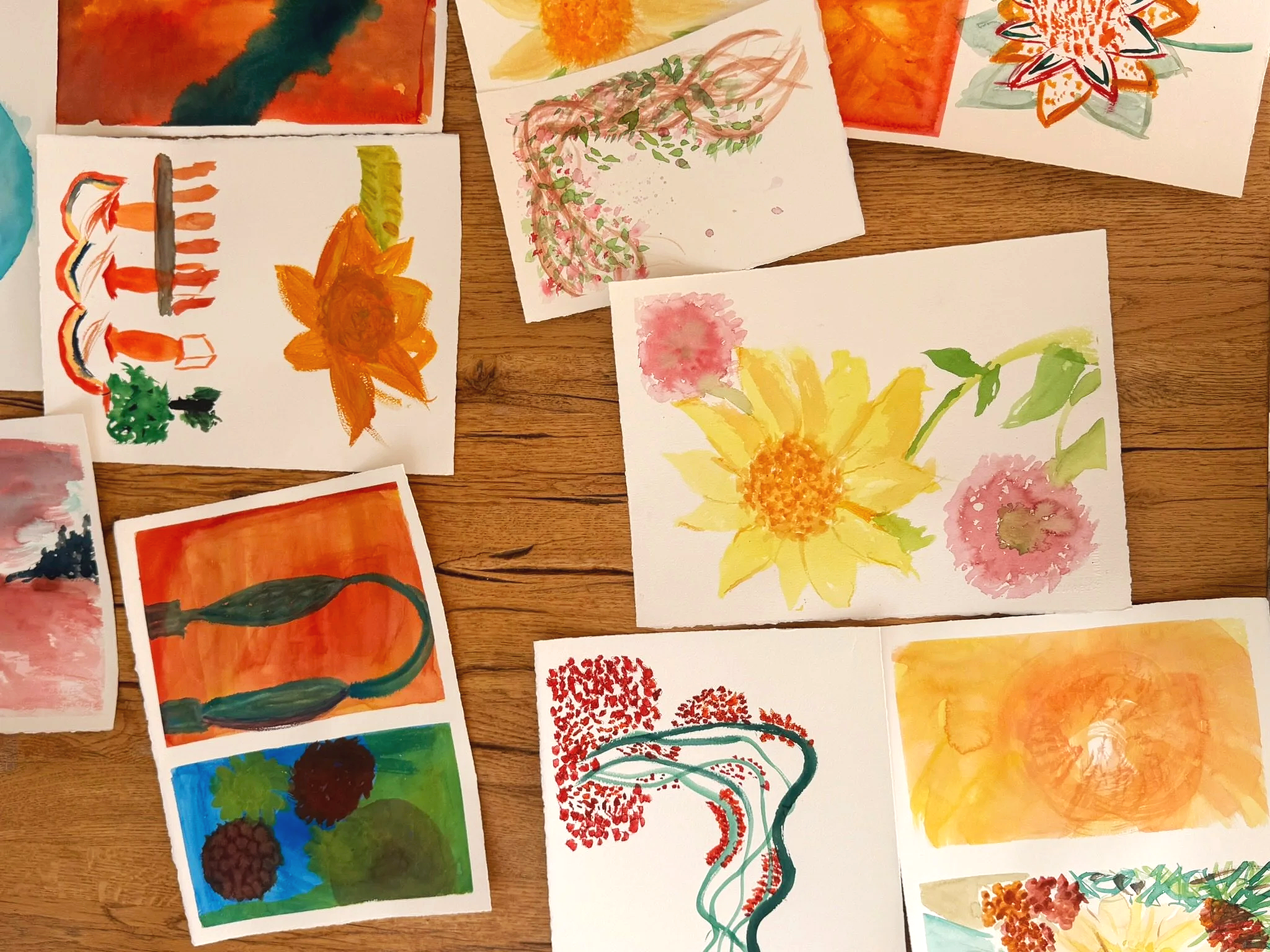 Watercolours from a 2026 summer art class at Casa Fuzetta in the Algarve; sunflowers, pink blooms, a flowering arch and bold orange washes, laid out on a wooden table.