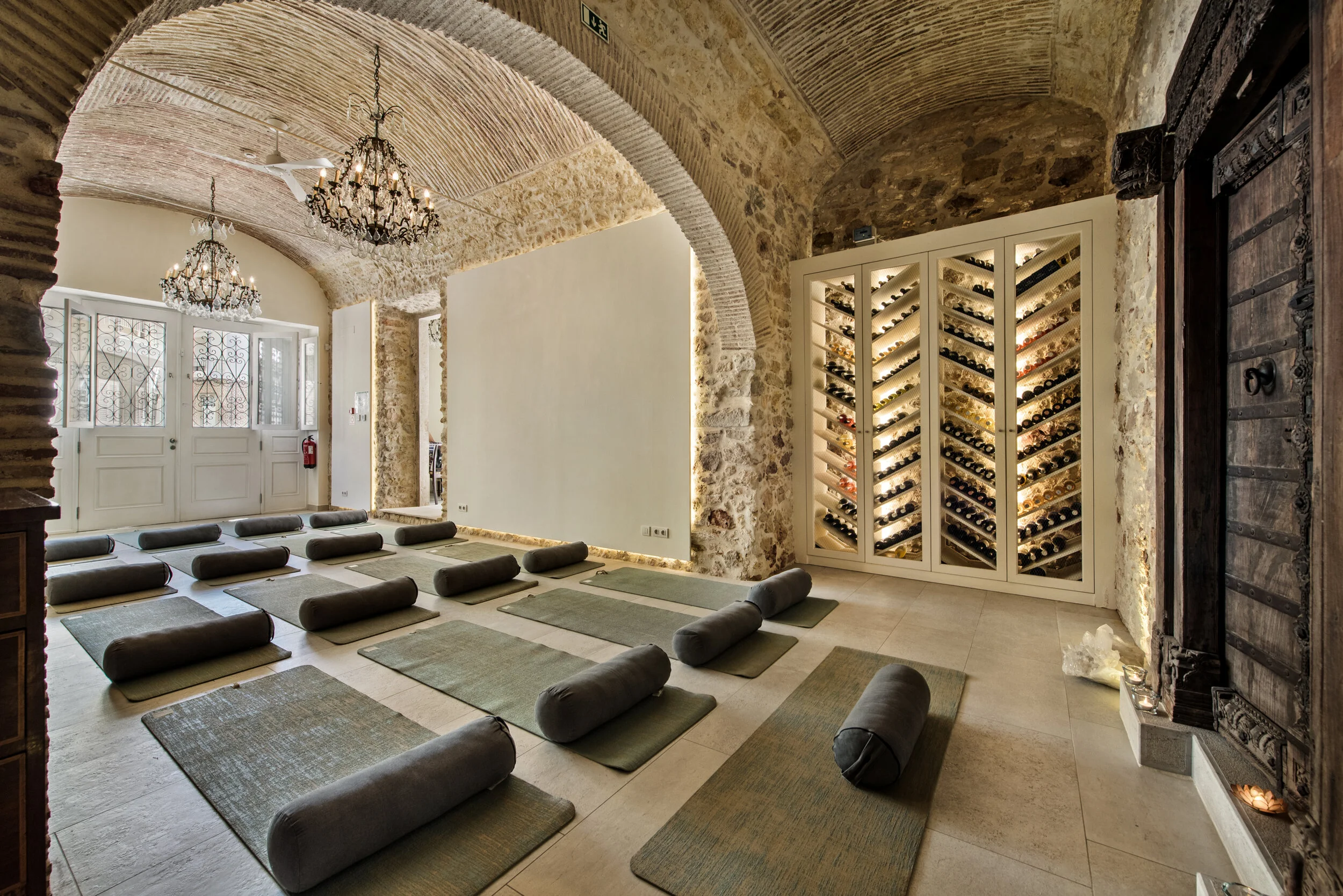 Image of the Casa Fuzetta stone walled entrance hall set up with yoga mats and boltsters for a class for 16 guests and one teacher also showing the backlit full wine cellar and the large overhanging chandeliers