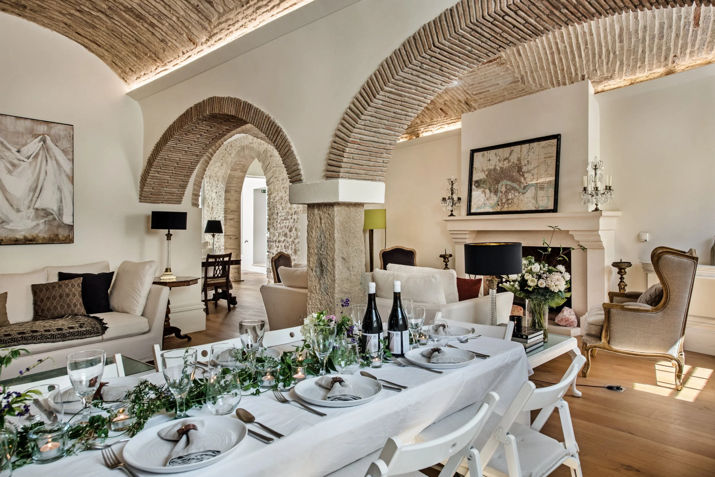 The living and dining room at Casa Fuzetta — a long table set with a herb-garland runner beneath a brick-vaulted ceiling and arches, with linen sofas and a stone fireplace.