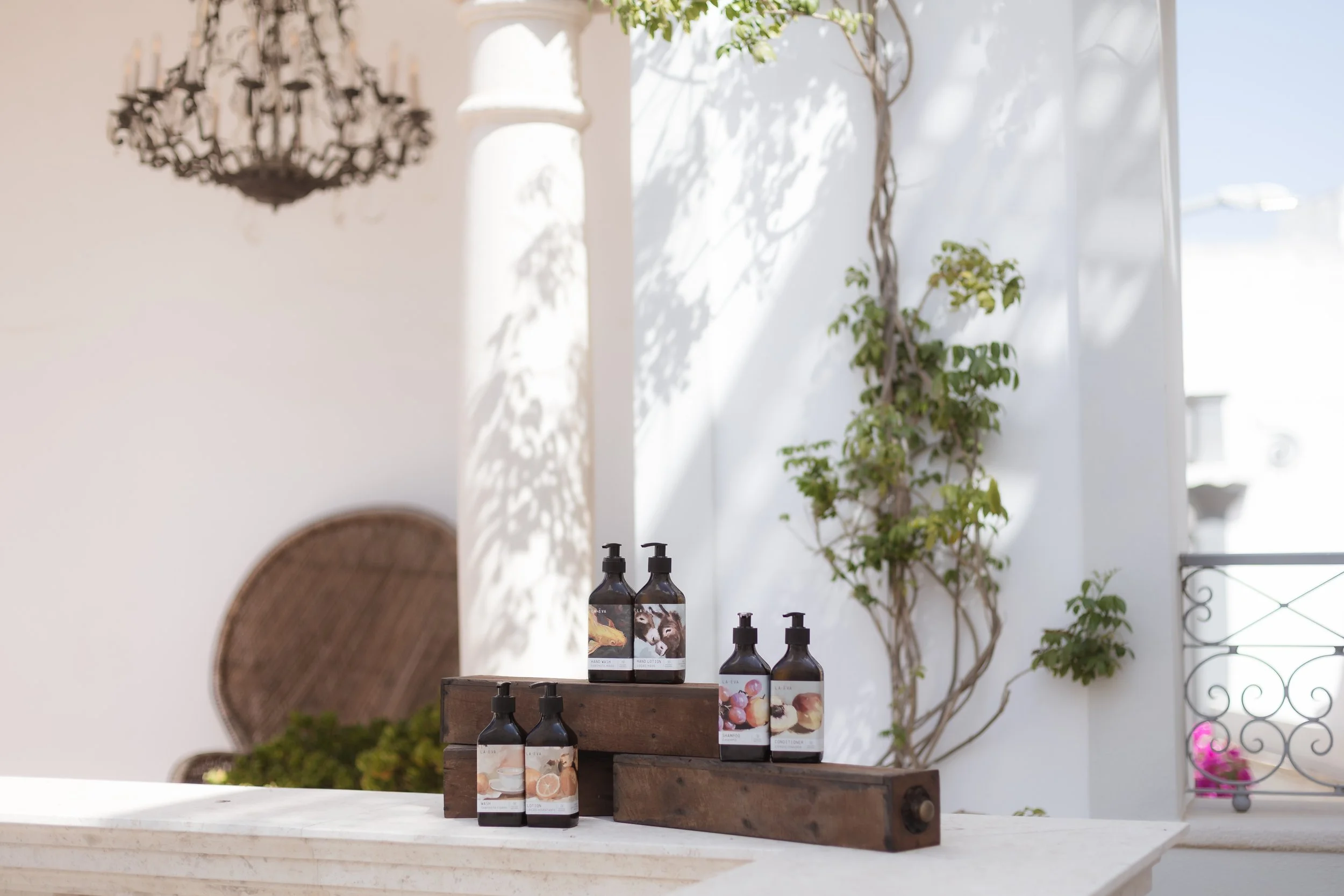 La-Eva Oro range bathroom amenities at Casa Fuzetta — illustrated amber pump bottles of hand wash, lotion, shampoo and conditioner on rustic wooden plinths in sunlit shade.