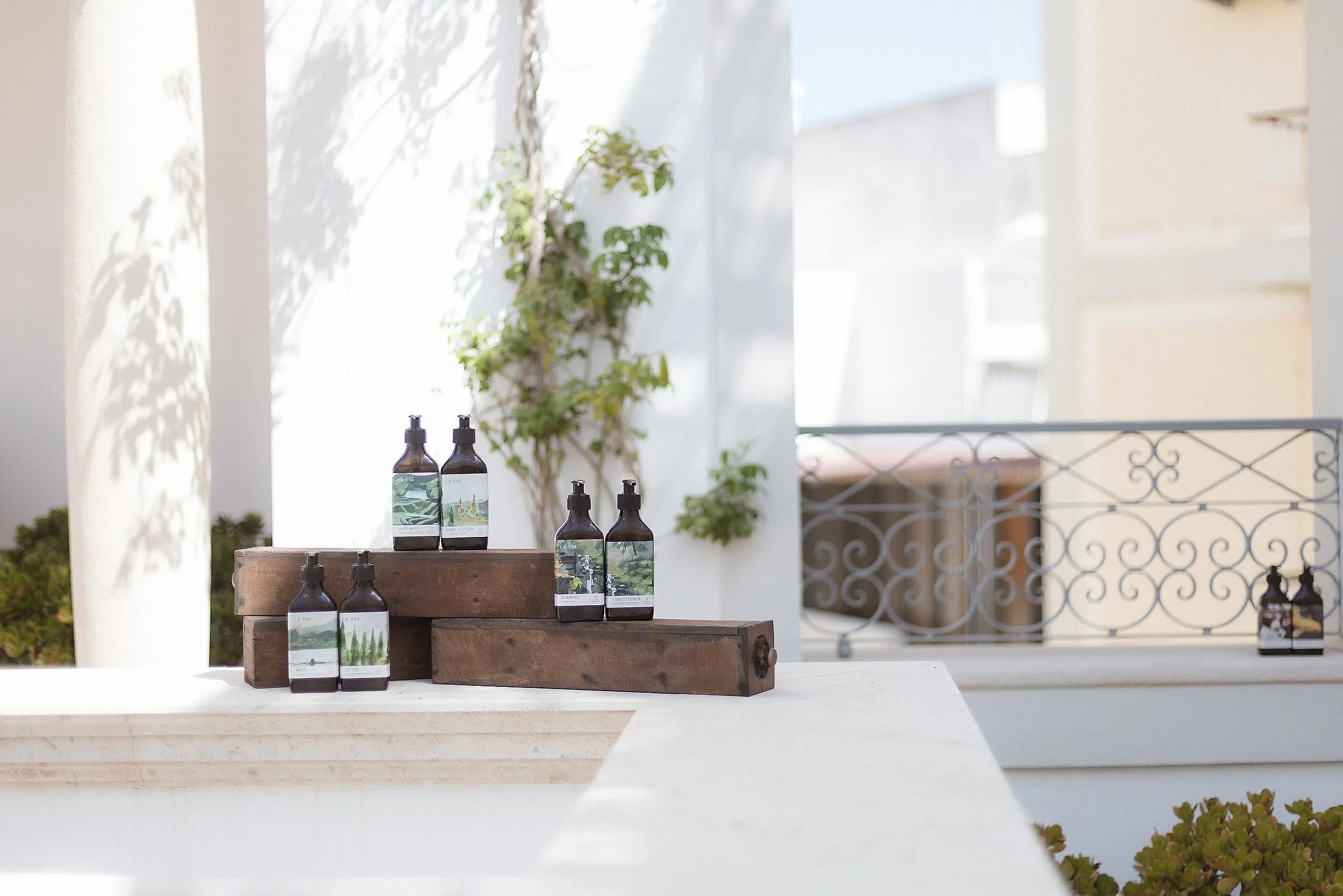 La-Eva Verte range bathroom amenities at Casa Fuzetta — amber pump bottles with green landscape labels arranged on rustic wooden plinths on a sunlit terrace.