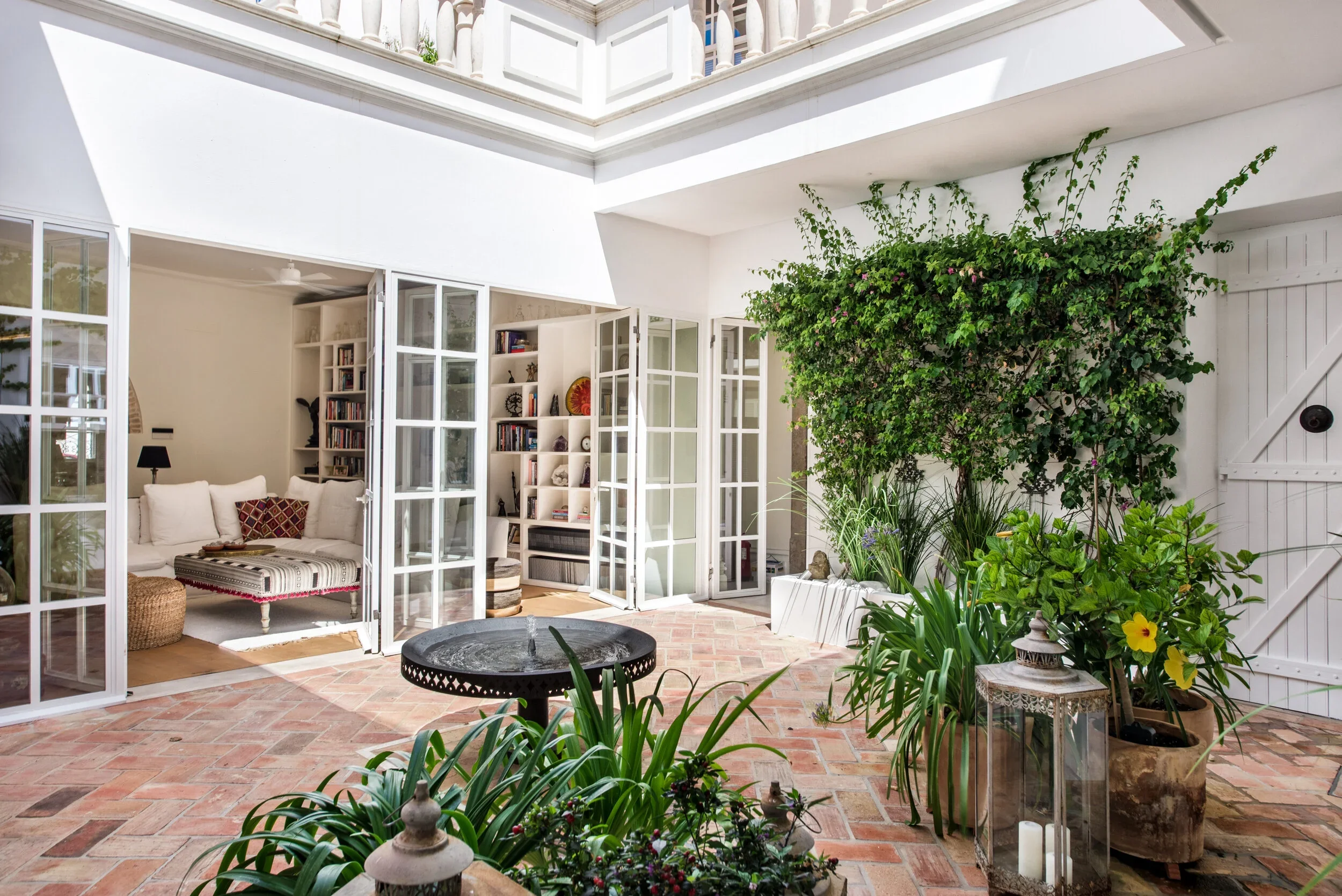 The planted inner courtyard at Casa Fuzetta in Olhão — terracotta-tiled and open to the sky, with a climbing fig, potted greenery, Moroccan lanterns and glazed doors opening to a library.