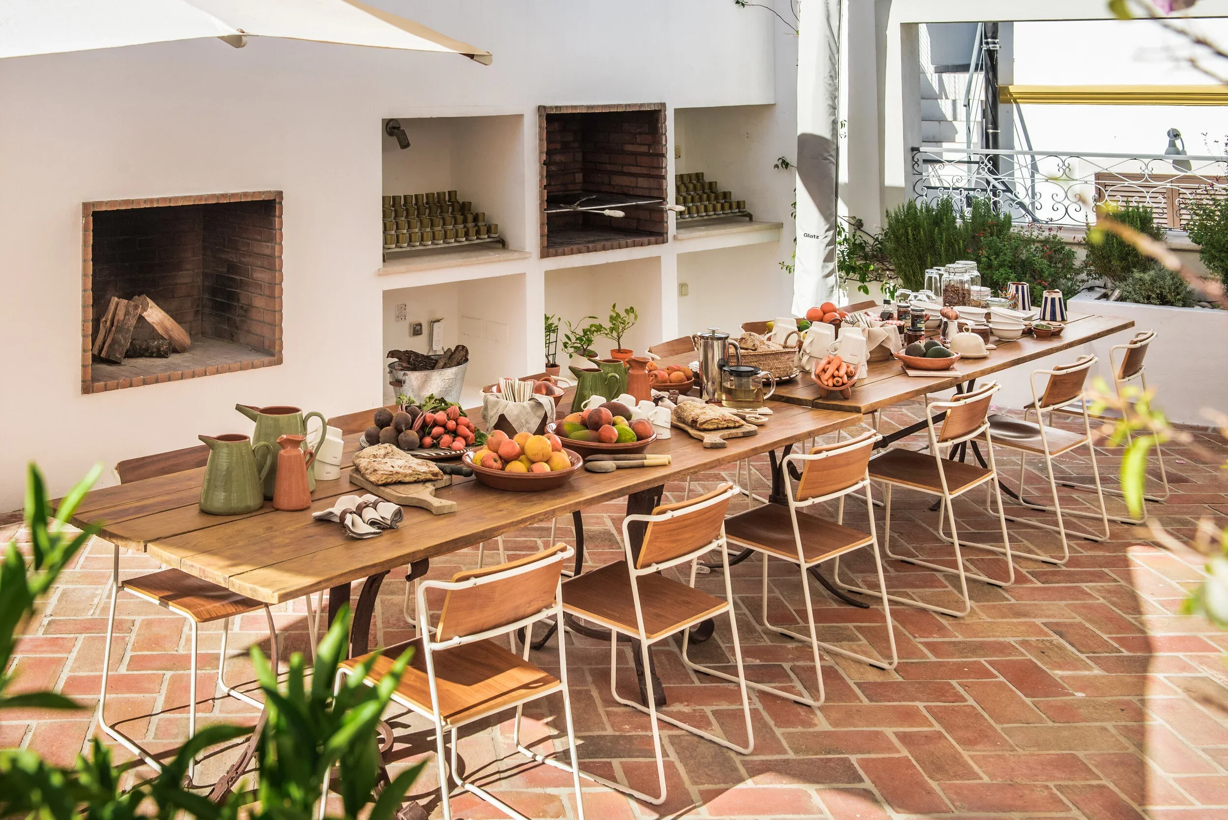 The kitchen terrace at Casa Fuzetta in Olhão laid for an alfresco feast — a table set with fruit, breads and ceramics, beside a barbecue and wood store, framed by potted herbs over terracotta tiles.