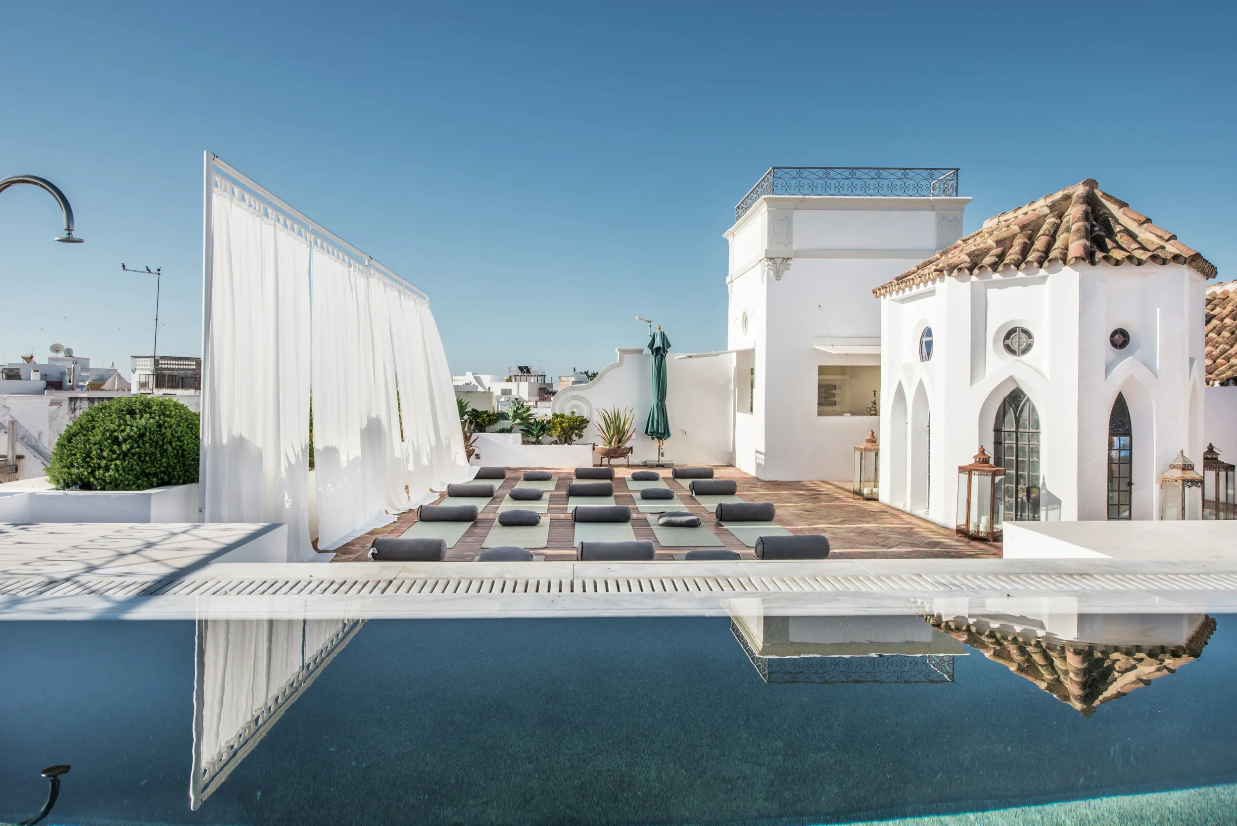The rooftop yoga deck at Casa Fuzetta — mats and bolsters on a tiled terrace beside a reflecting pool and a whitewashed Moorish-arched pavilion, under a clear blue sky.