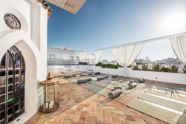 The rooftop yoga deck at Casa Fuzetta in Olhão — mats and bolsters on a terracotta terrace beside an arched pavilion with a stained-glass window, white curtains and a brass lantern, under blue sky.