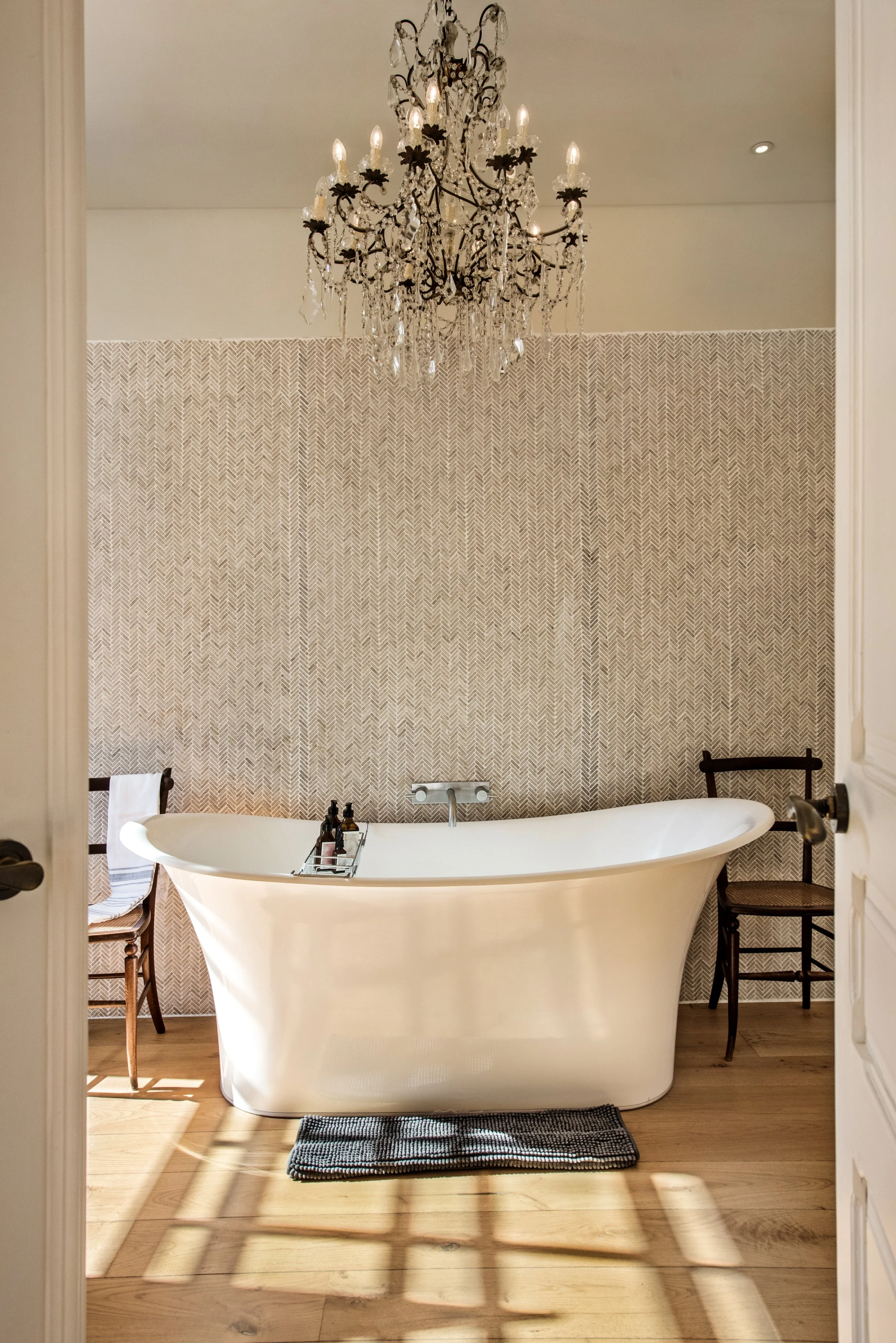 The ensuite bathroom of the Crystal Suite at Casa Fuzetta in Olhão, with a freestanding roll-top bath against a herringbone tiled wall, a crystal chandelier and oak floors washed in afternoon light.
