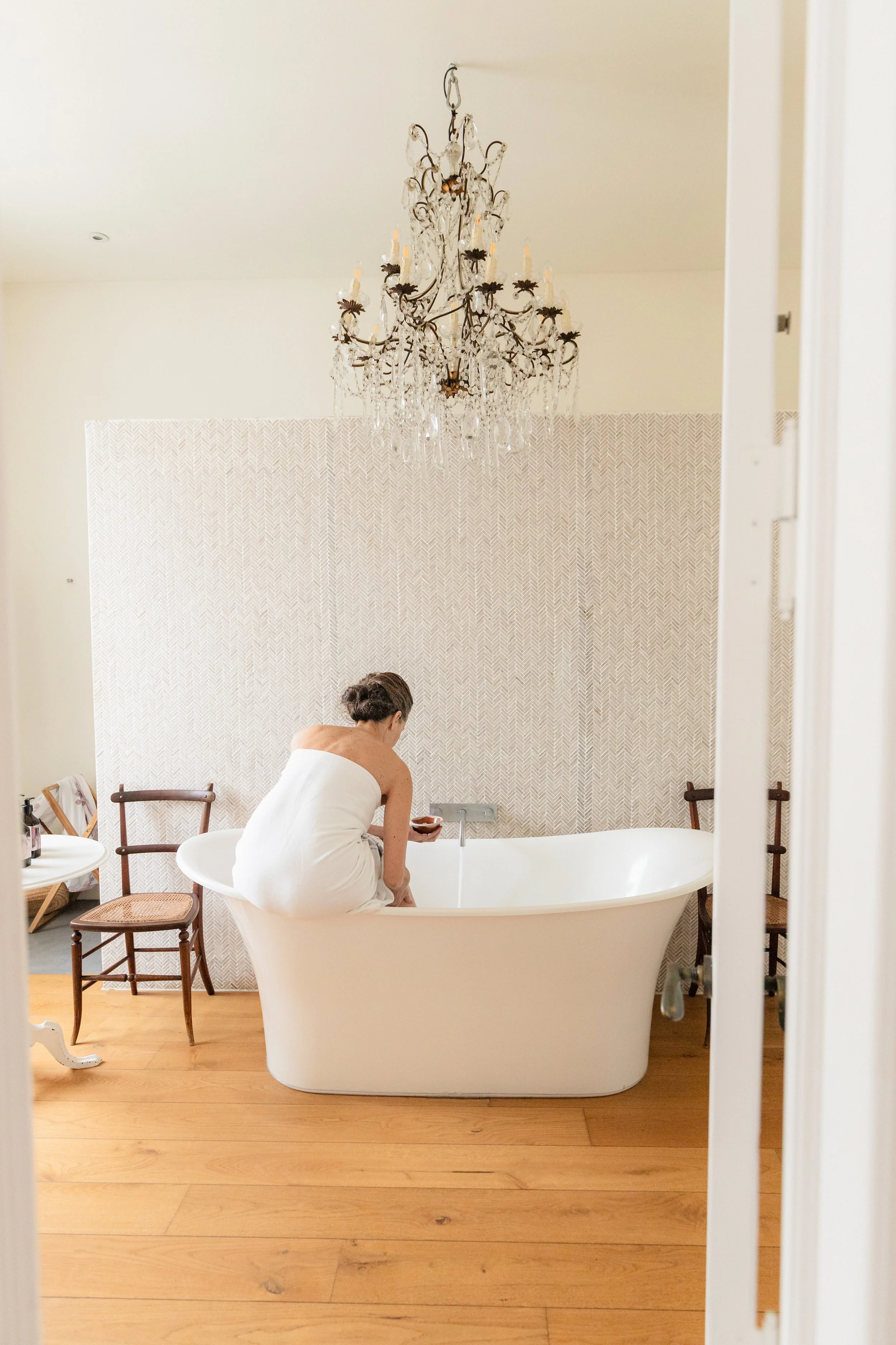 A woman in a towel adding bath salts to a freestanding roll-top bath beneath a crystal chandelier, against a herringbone-tiled wall, in the Crystal Suite at Casa Fuzetta in Olhão.