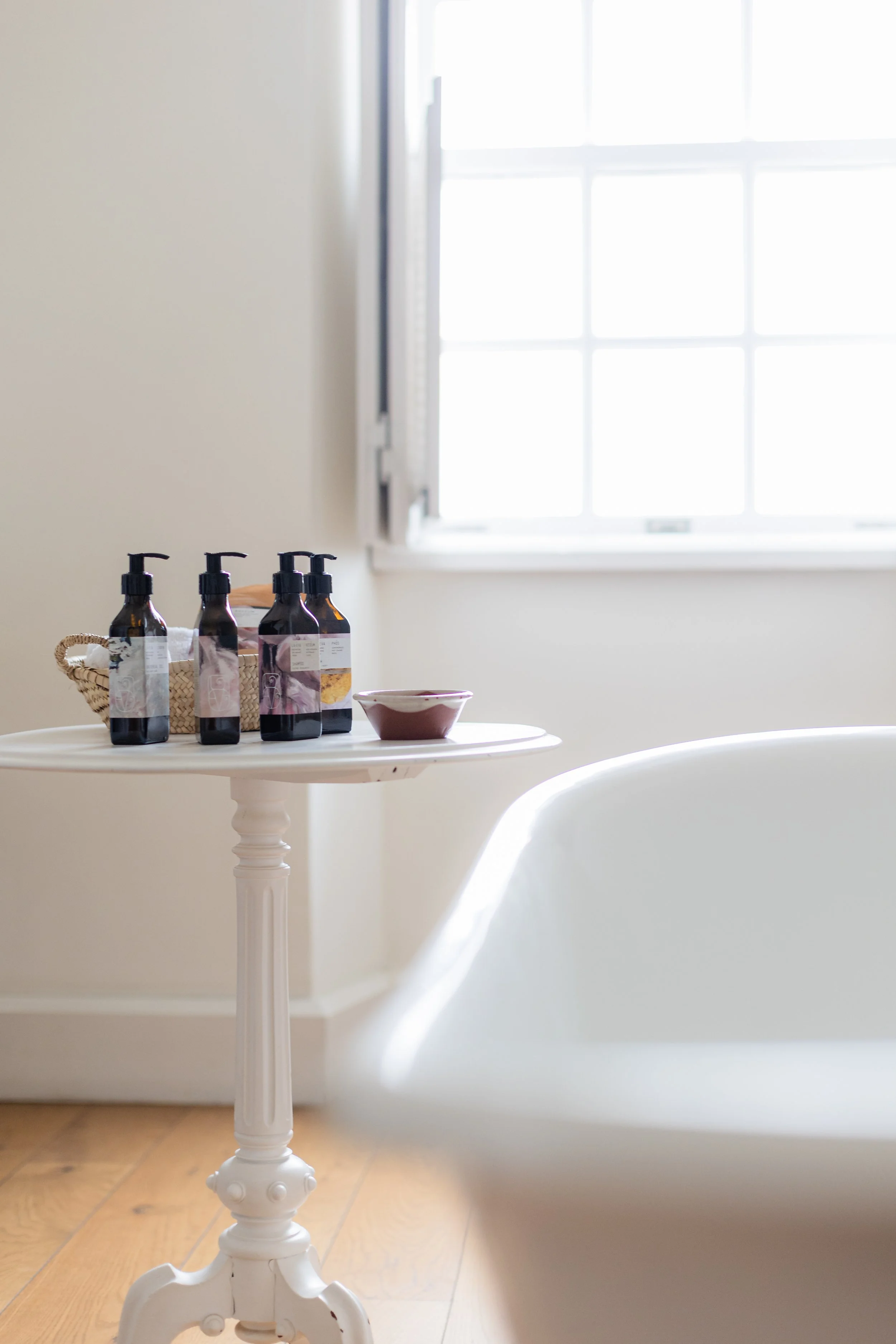 A woven basket of La-Eva amber-bottle amenities and a ceramic dish on a marble pedestal table beside a roll-top bath, in a light-filled bathroom at Casa Fuzetta in Olhão.