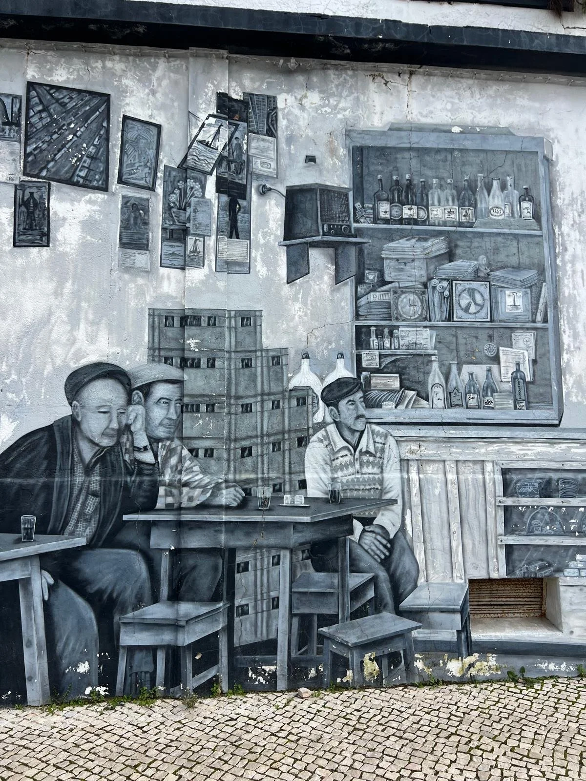 A large black-and-white mural in Olhão's old canning district, a short walk from Casa Fuzetta, showing three older men in flat caps drinking around a table in a traditional Portuguese taberna.