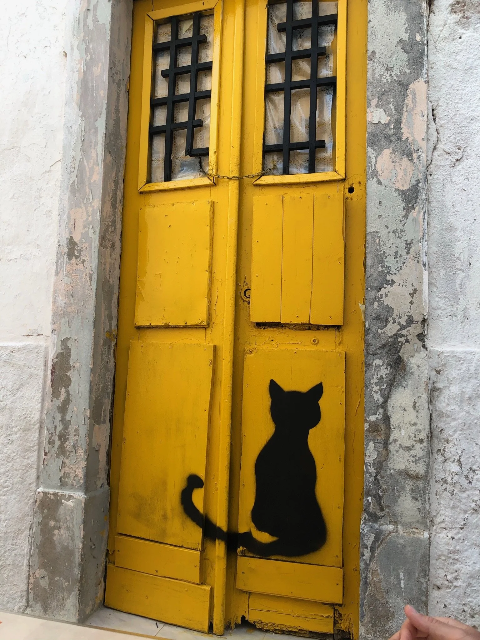 A weathered mustard-yellow double door with a black cat stencilled on the lower panel, set into a peeling whitewashed wall on a narrow street in Olhão, close to Casa Fuzetta.