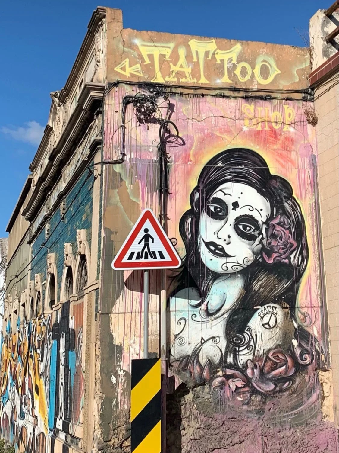 A large street art mural on a tattoo shop corner in Olhão, close to Casa Fuzetta; a woman's face in Day of the Dead sugar-skull style, with dark hair, roses and a peace symbol tattoo.