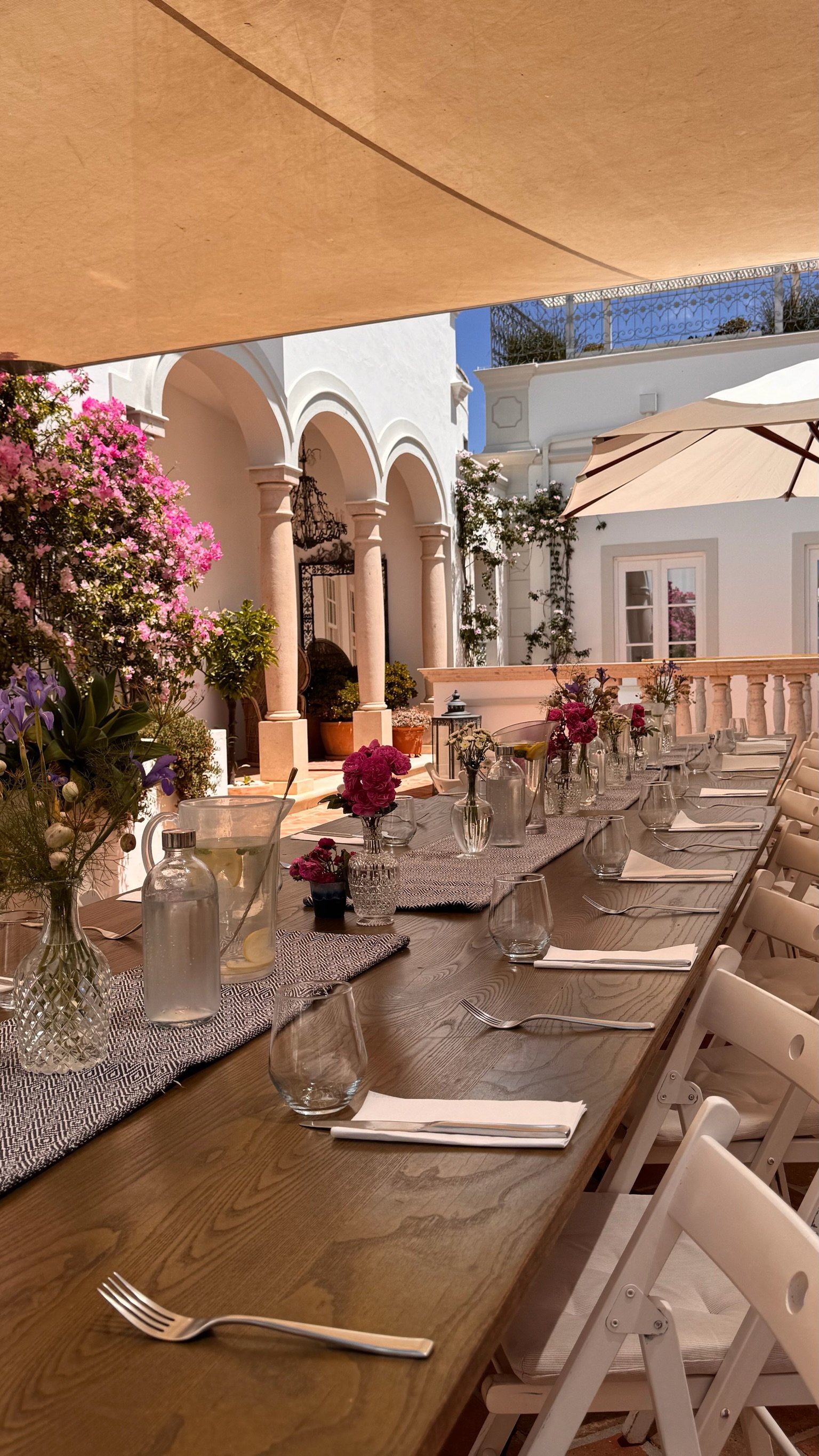 A long wooden dining table set for lunch under a canopy in Casa Fuzetta's collonaded courtyard in Olhão, with pitchers of iced water, wildflowers and pink bougainvillaea by the stone columns.
