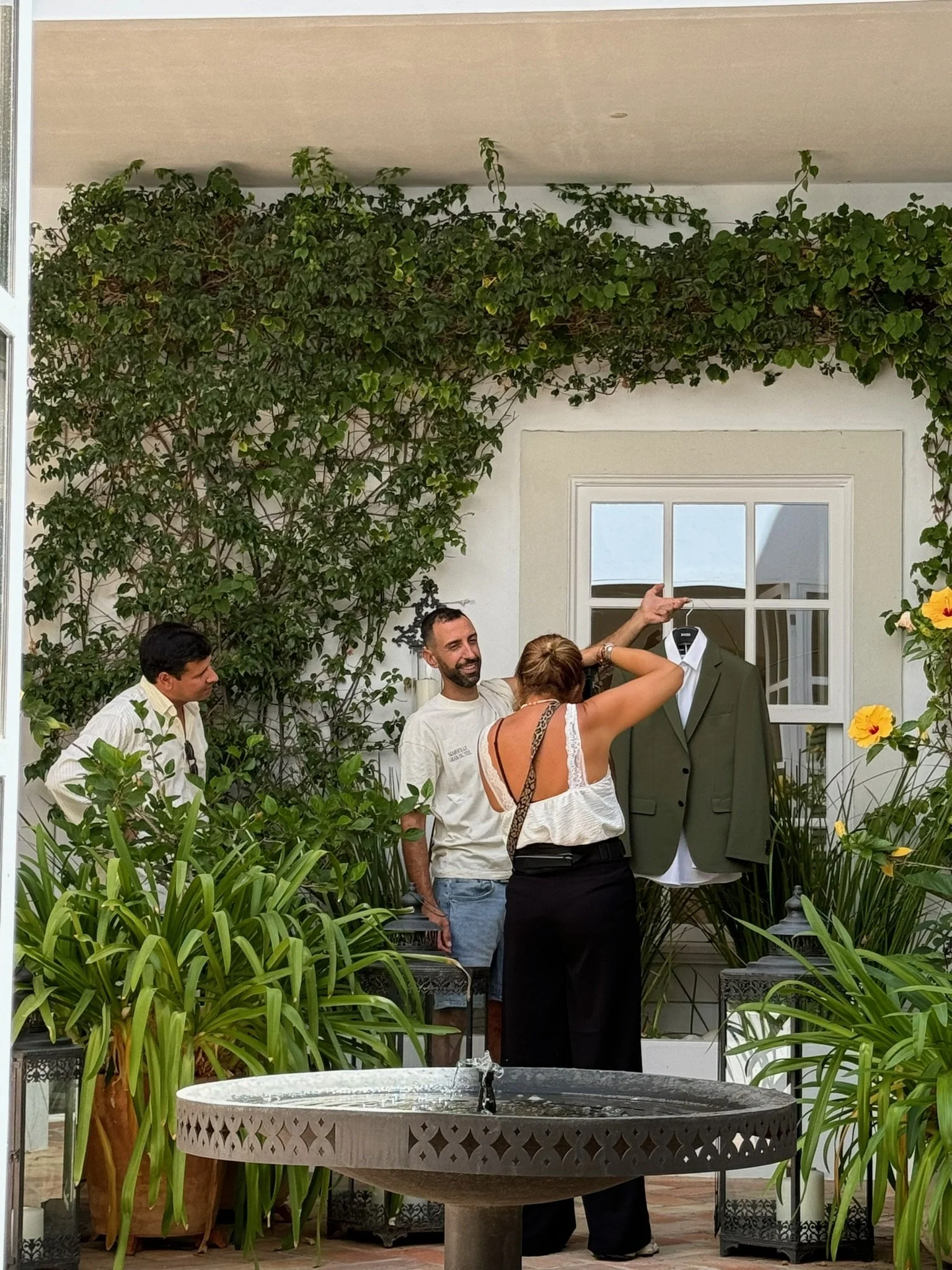 Wedding preparations at Casa Fuzetta in Olhão. A woman holds up an olive green suit jacket by the ivy-clad courtyard fountain as two men look on, hibiscus in bloom nearby.
