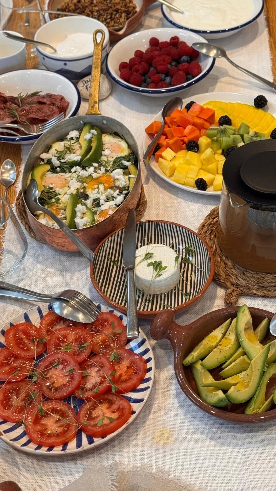 A breakfast spread on white linen at Casa Fuzetta; a copper pan of baked eggs with avocado and feta, fresh cheese with herbs, sliced tomato, granola, yogurt, berries and tropical fruit.
