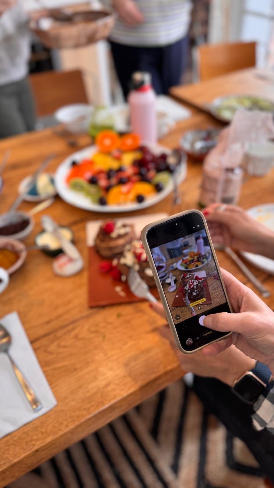 A guest photographs a granola pot on the breakfast table at Casa Fuzetta on their phone, a fruit platter and other dishes spread around them.