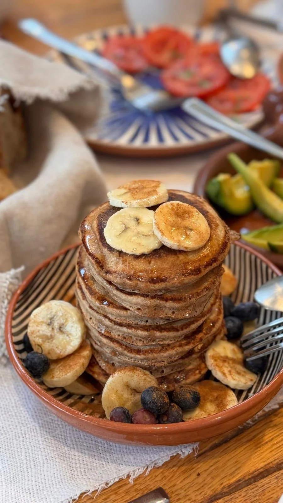A stack of banana pancakes with cinnamon and blueberries in a terracotta dish at Casa Fuzetta, with sliced tomatoes and avocado on the breakfast table behind.