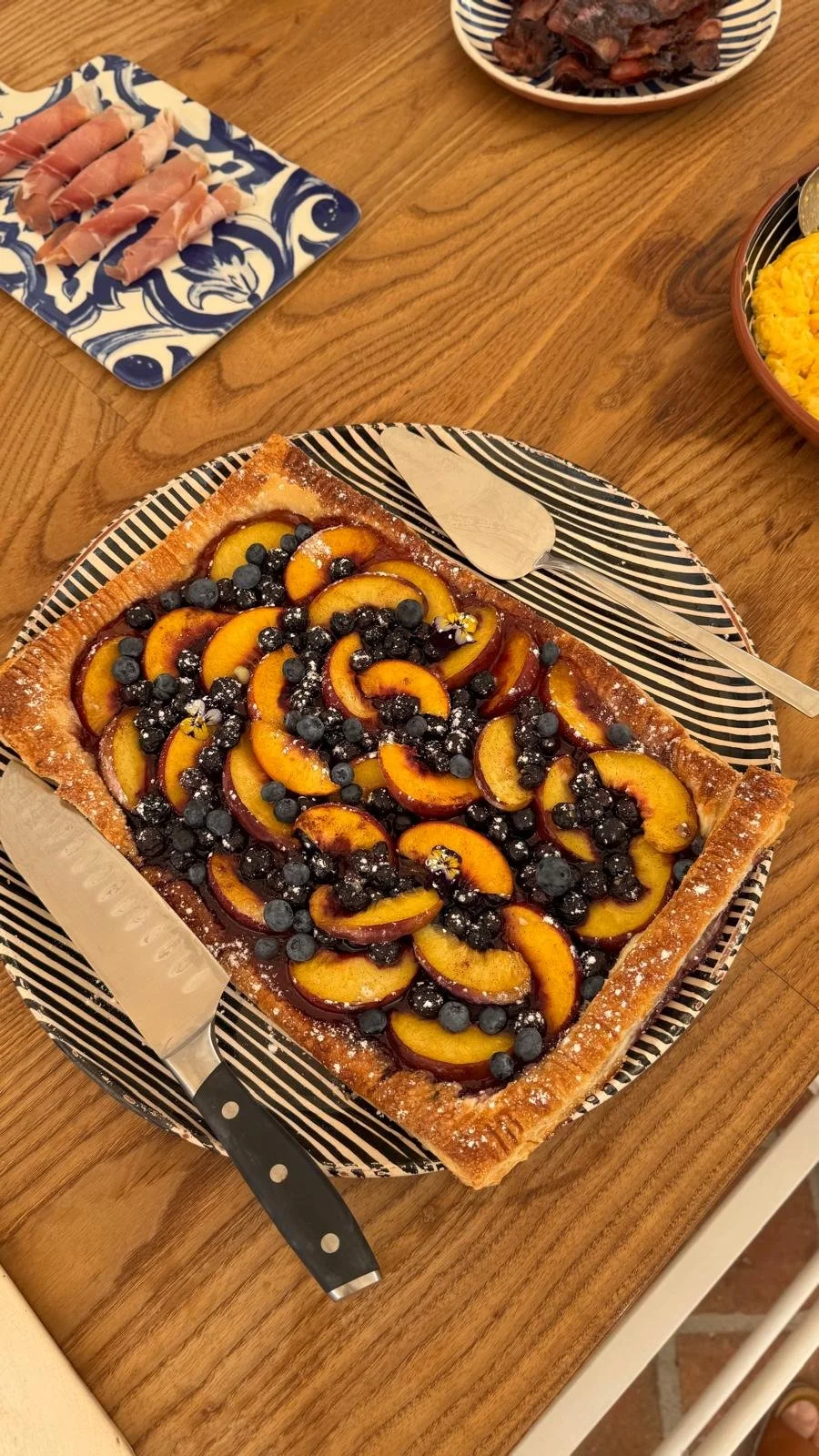 A rustic peach and blueberry puff-pastry tart dusted with icing sugar on a striped platter at Casa Fuzetta, with rolls of prosciutto plated on blue azulejo tiles alongside.