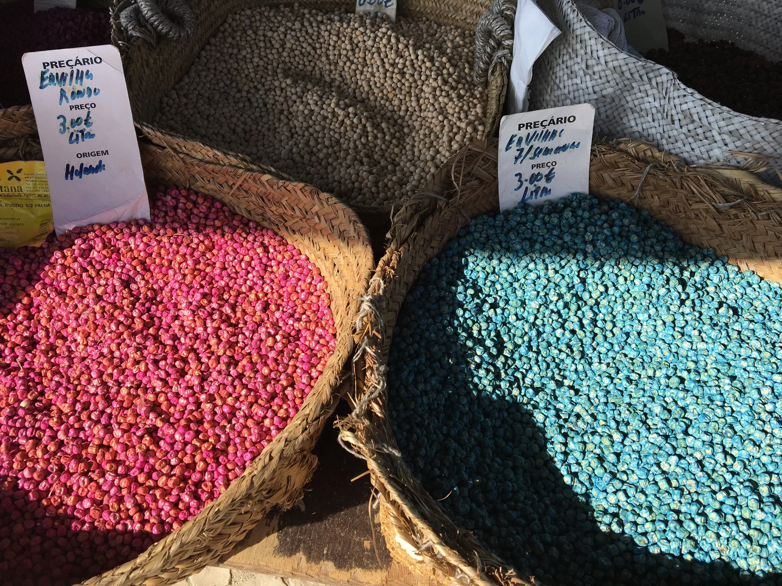 Wicker baskets of dyed seed peas at the Mercado de Olhão; the pink and turquoise coatings mark them as pea seeds for planting, price cards hand-written in Portuguese.