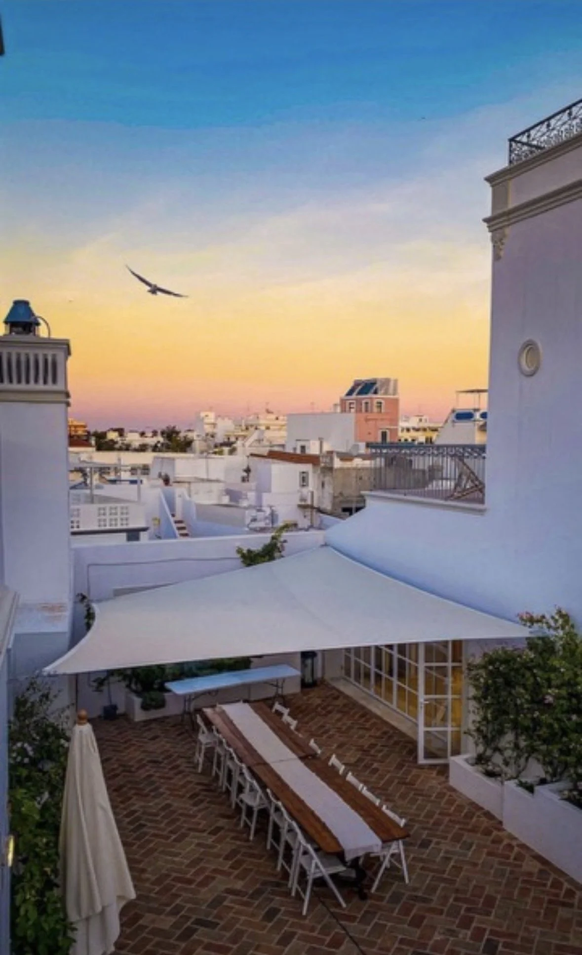 The kitchen terrace at Casa Fuzetta at sunset; a long wooden dining table beneath a white sail canopy on herringbone brick, a gull passing above Olhao’s whitewashed rooftops to the horizon.