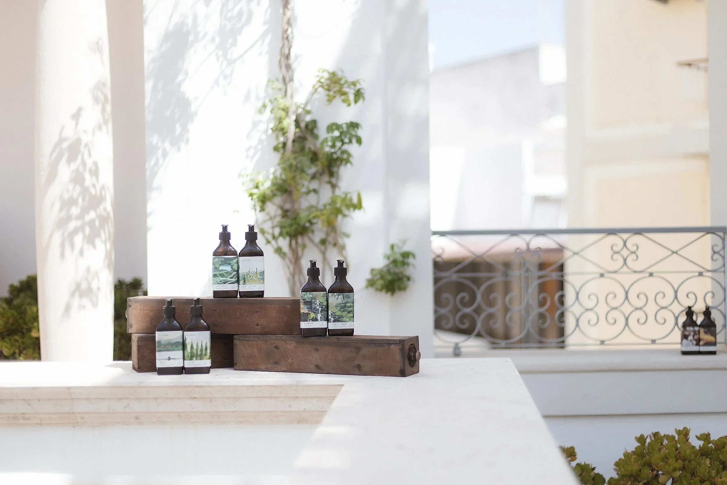 The full La-Eva Verte range of wash, lotion, shampoo, conditioner and hand care in amber bottles with green landscape labels, arranged on weathered timber blocks on a sunlit terrace at Casa Fuzetta in Olhão.