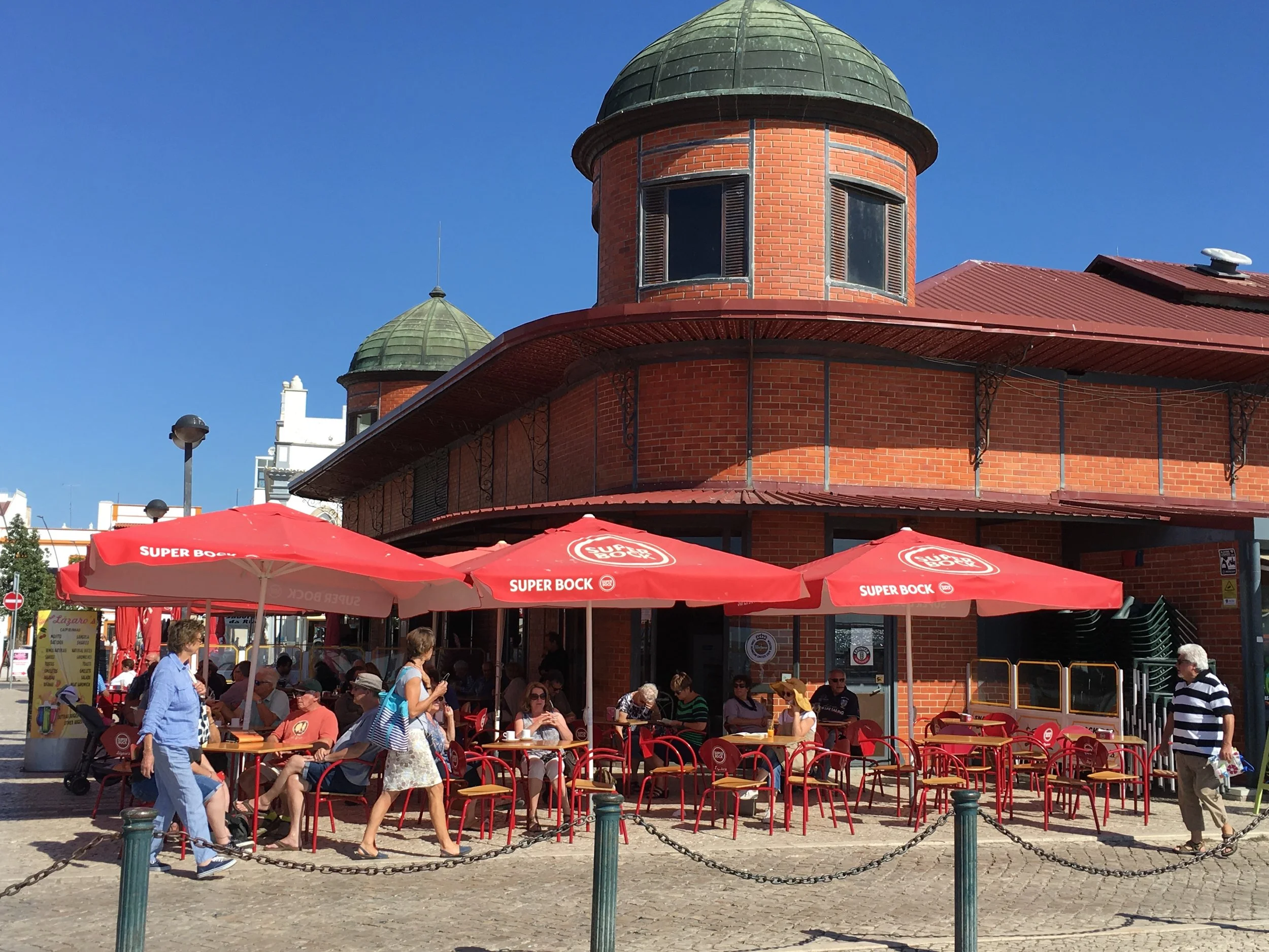 The red-brick Mercado de Olhão with its domed towers under a clear blue sky, a short walk from Casa Fuzetta, with people at red café tables and parasols on the cobbled square outside.