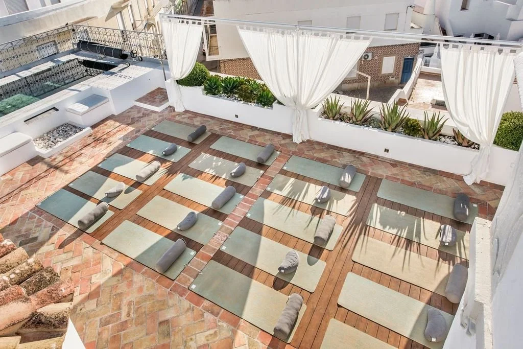 The rooftop yoga deck at Casa Fuzetta seen from above; rows of sage mats and grey bolsters on terracotta and timber, with white curtains, planters and a pool, over the Olhao rooftops.