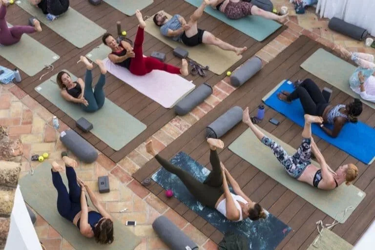 An overhead view of a Cheeky Yoga Club Pilates class on the Casa Fuzetta rooftop in Olhão, participants on mats with bolsters and water bottles, on wooden decking edged in terracotta.