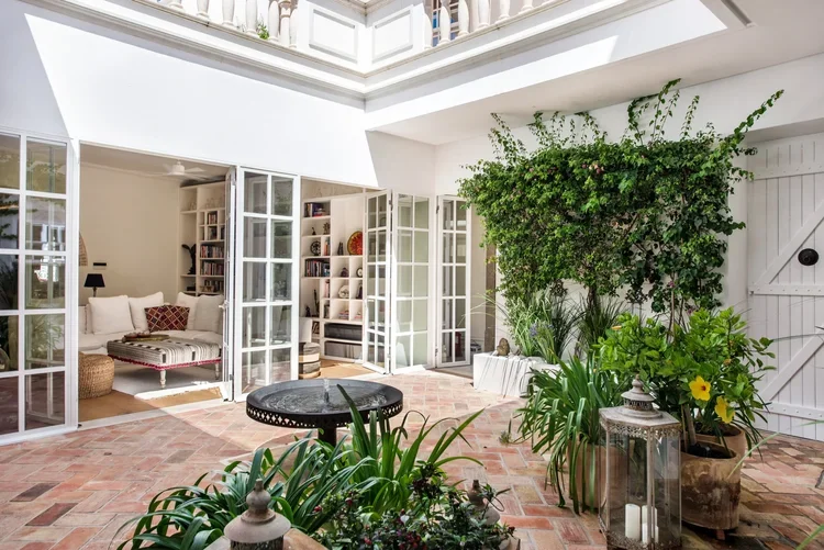 The interior courtyard at Casa Fuzetta; a round black fountain on a terracotta-tiled floor amid potted plants and climbing bougainvillea, with glass doors opening to a book-lined sitting room.