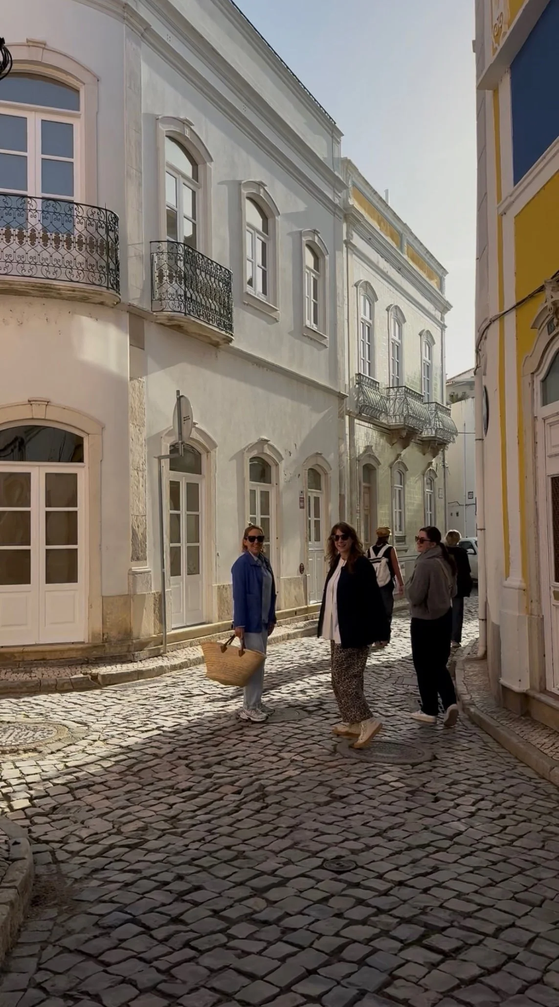 Three women pause on a sunlit cobbled street in Olhão, close to Casa Fuzetta, among whitewashed townhouses with wrought-iron balconies, one carrying a woven straw basket.