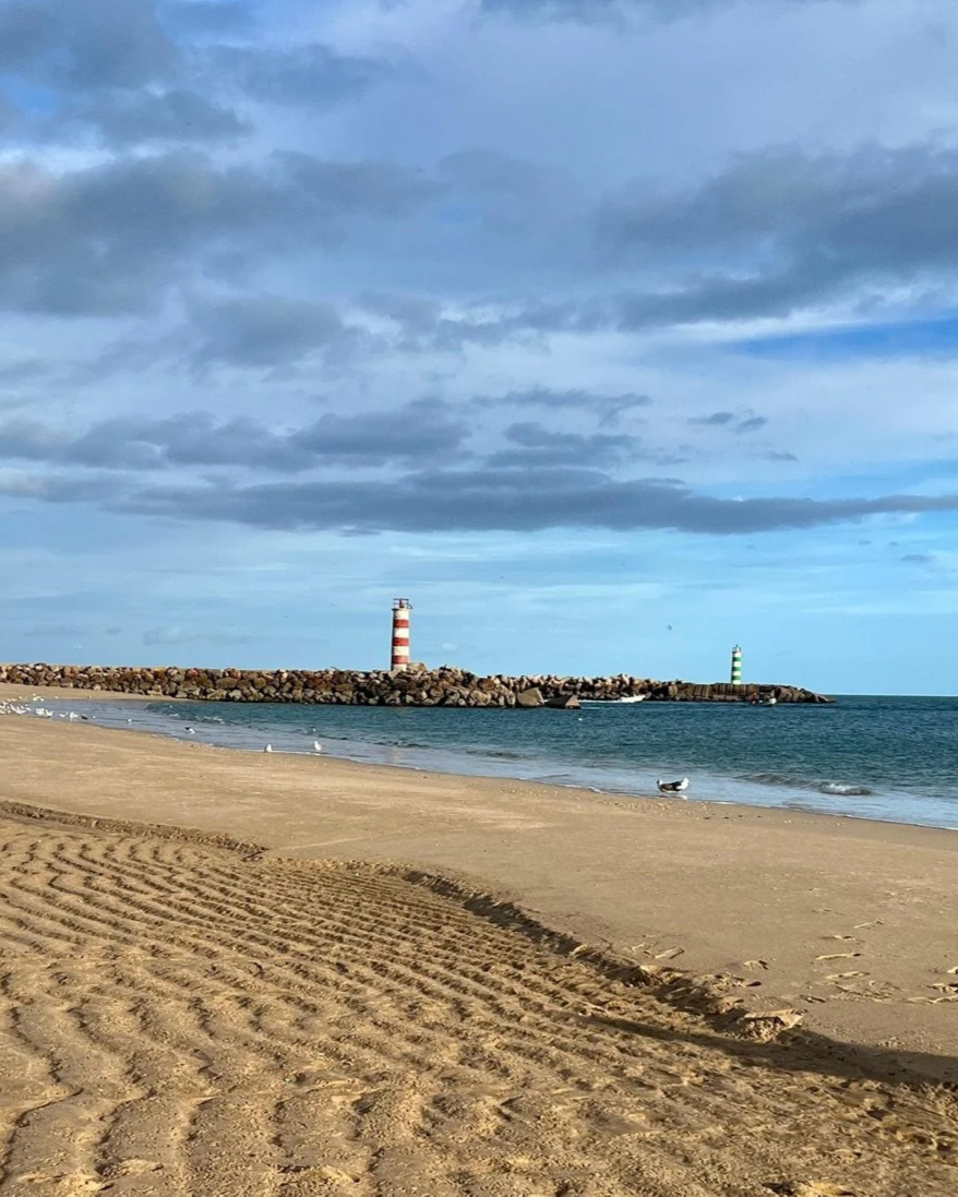 The empty rippled-sand beach at Ilha Deserta on the Ria Formosa, a short river-taxi from Casa Fuzetta, with two striped lighthouses on a rocky breakwater under a cloudy sky.