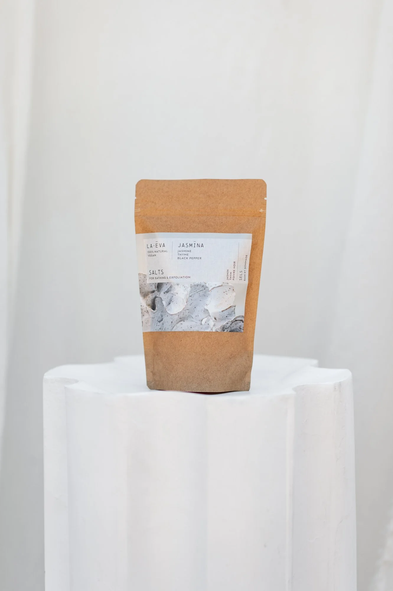La-Eva Jasmina Exfoliant & Bath Salts in a kraft pouch — scented with jasmine, thyme and black pepper — displayed on a white fluted plinth
