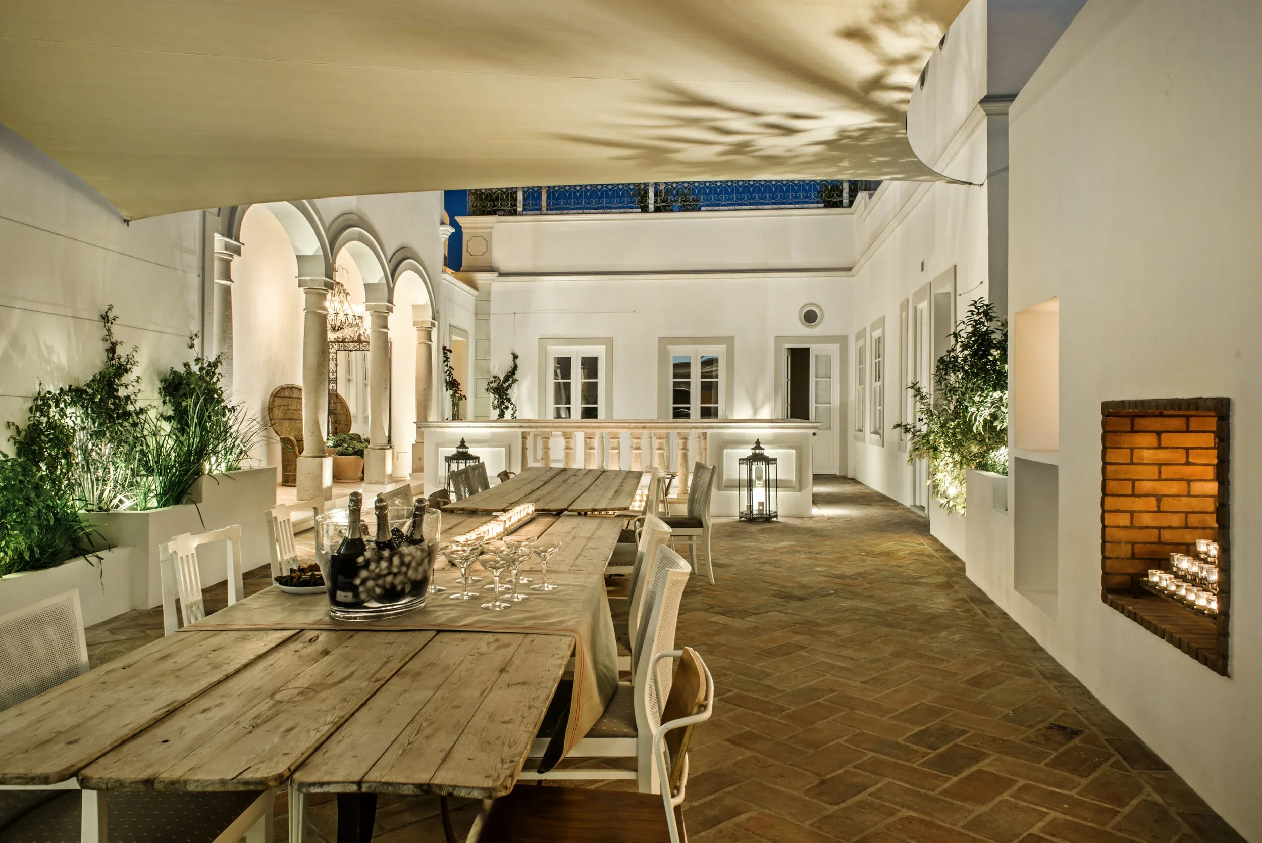 The kitchen terrace at Casa Fuzetta at dusk; a long reclaimed-wood table with champagne on ice and coupe glasses, beside an arched stone colonnade and candlelit niches.