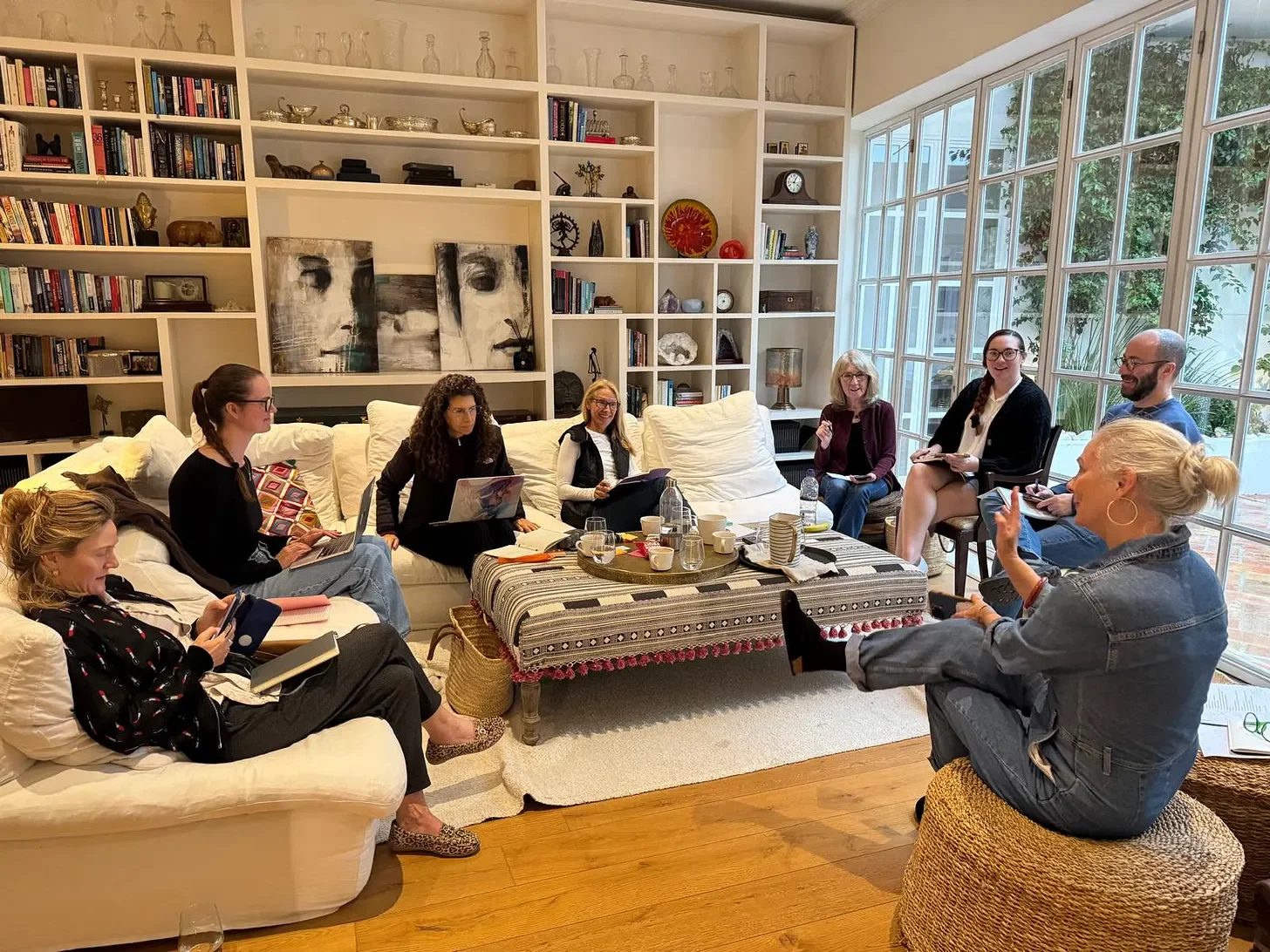 A writing retreat hosted by Jacq Burns, founder of the London Writers' Club, in the library at Casa Fuzetta — guests sit talking on white sofas around a patterned ottoman in a book-lined room.