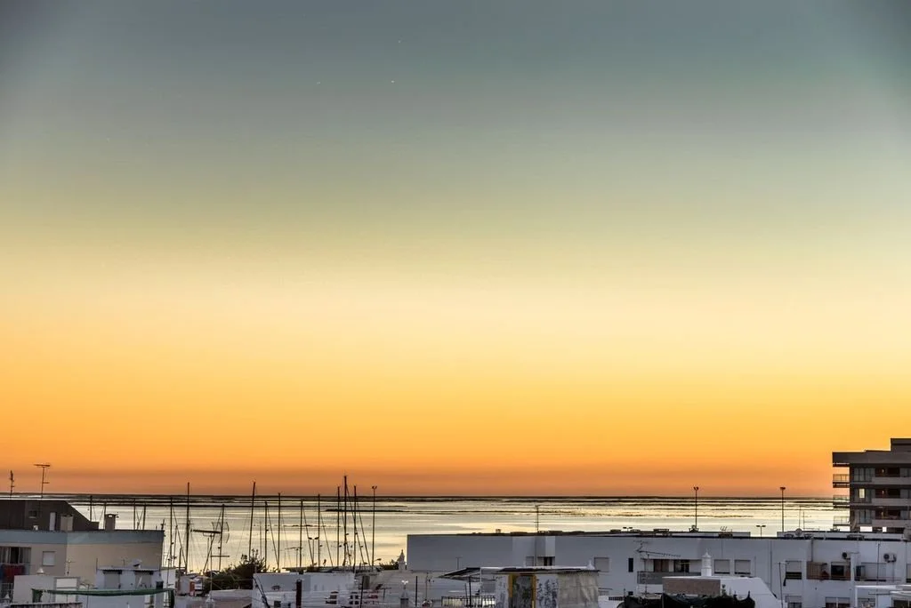 The view from Casa Fuzetta over Olhao and the Ria Formosa — a graduated teal-to-amber sky above the lagoon's sandbar islands, marina masts and the white rooftops of the old town.